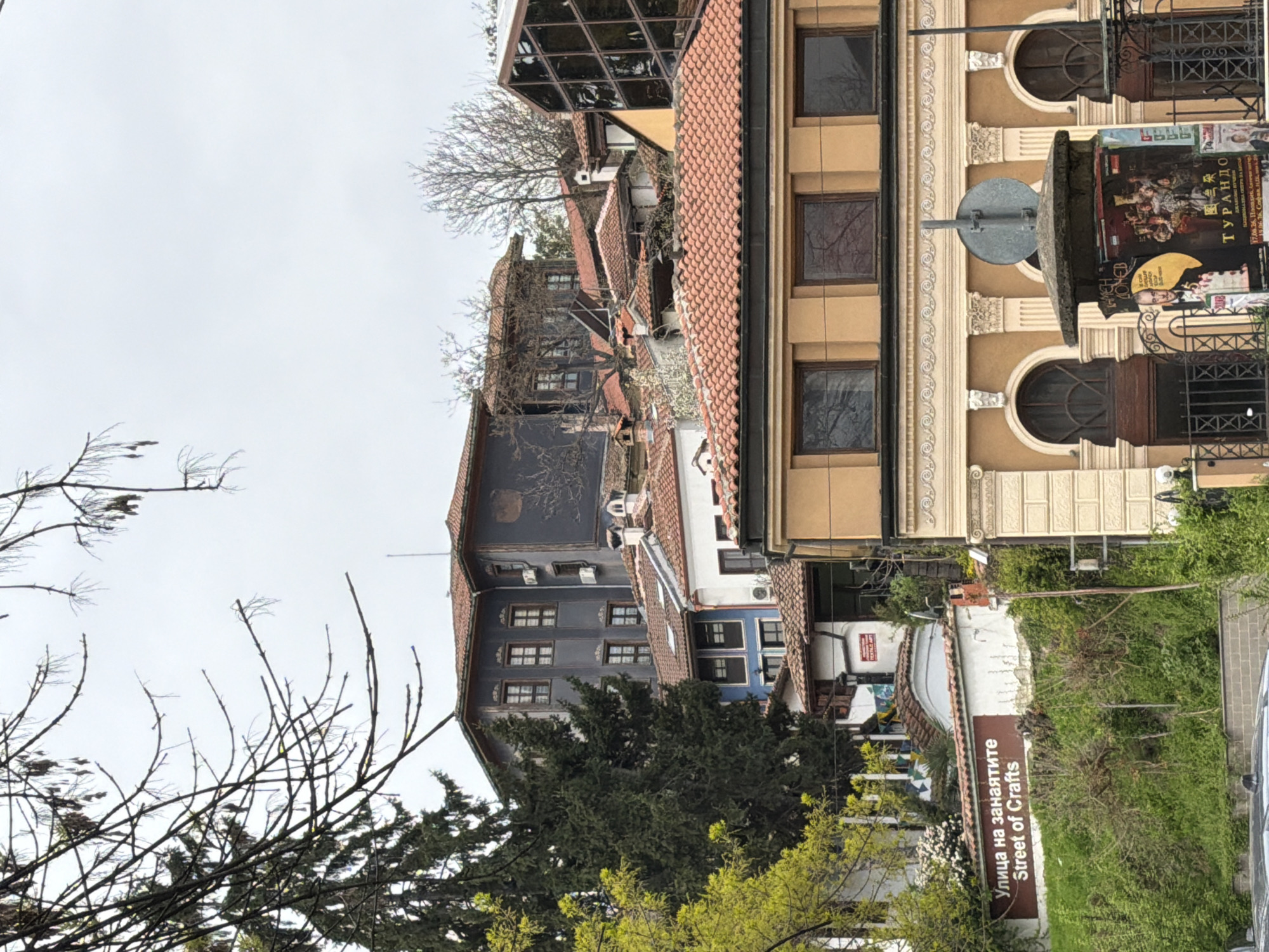 The image shows a street scene in Plovdiv, Bulgaria. On the right side of the image, there's a tall building with a beige facade. Its windows are rectangular and dark, and there is a circular architectural detail above each window on the top floor. A sign displaying posters is affixed to the building. Adjacent to the street is a sign that says "Улица на занаятите Street of Crafts," indicating the location. The street is lined with a low white wall overgrown with foliage. Further down the street, there are older buildings with darker facades and red-tiled roofs. Some trees are present, some with bare branches and some with budding green leaves. The sky is overcast.