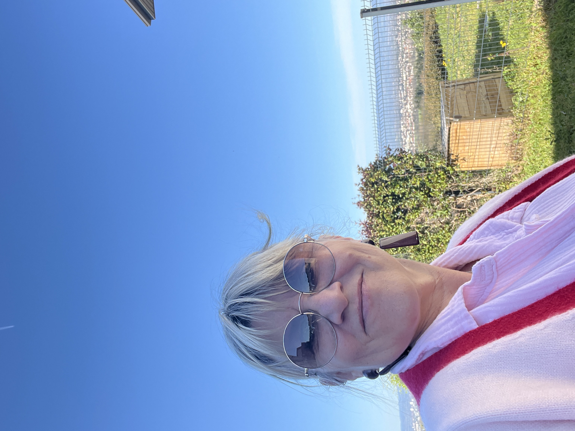 A woman with blonde hair and round sunglasses is photographed outdoors under a clear blue sky. She is wearing a pink and white striped sweater. The background includes green grass, bushes, and a wire fence, with buildings visible in the distance, possibly the town of Chambœuf, France.
