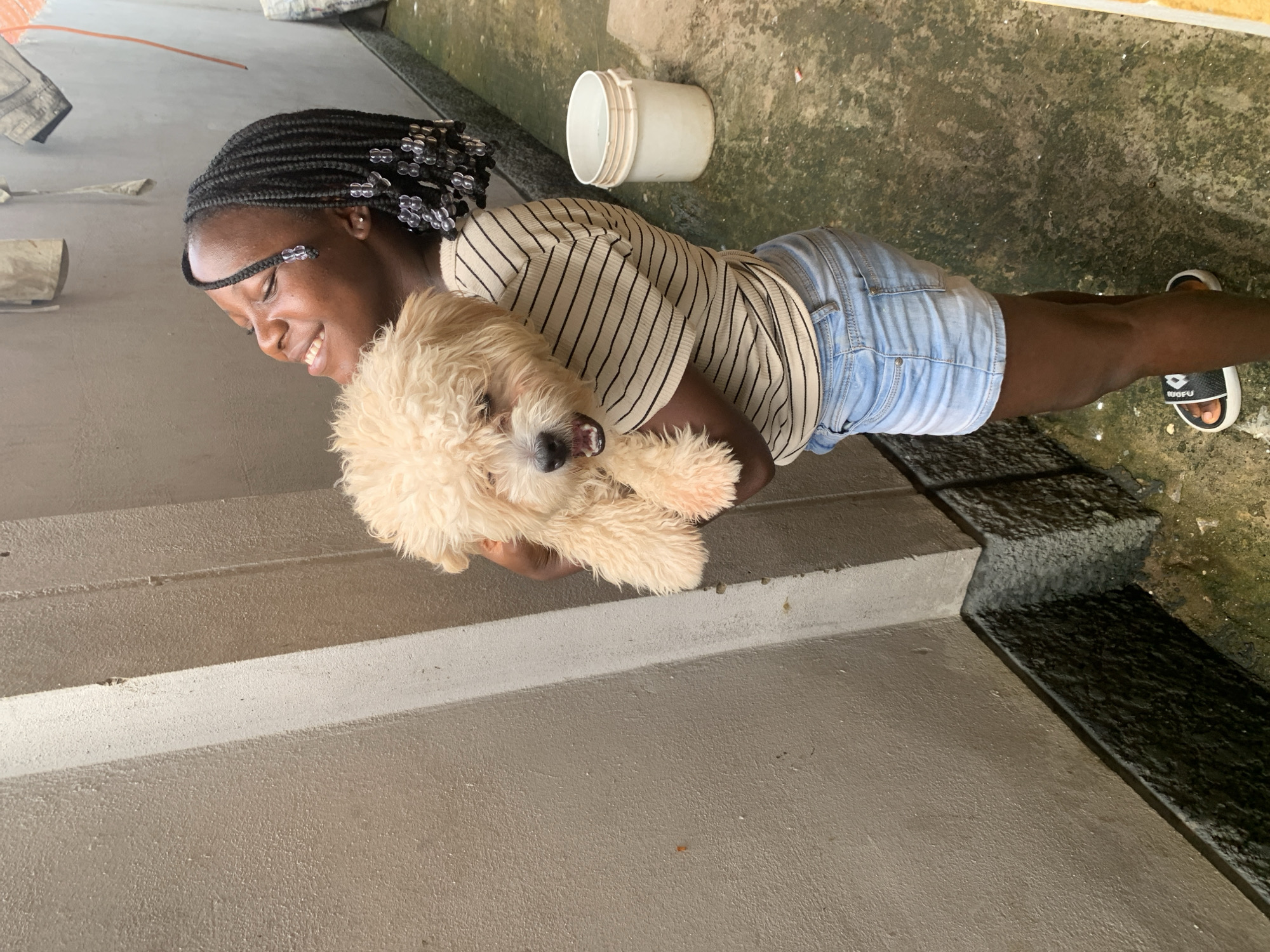A young Black female with braided hair, wearing a striped t-shirt and denim shorts, holds a fluffy cream-colored dog. She stands outdoors next to a textured concrete wall. Her eyes are closed and she is smiling, looking up. The dog has its mouth slightly open. To the right of the woman is a white bucket filled with water. The ground is concrete and appears wet and a bit dirty. The scene appears to be in an outdoor alleyway or courtyard.