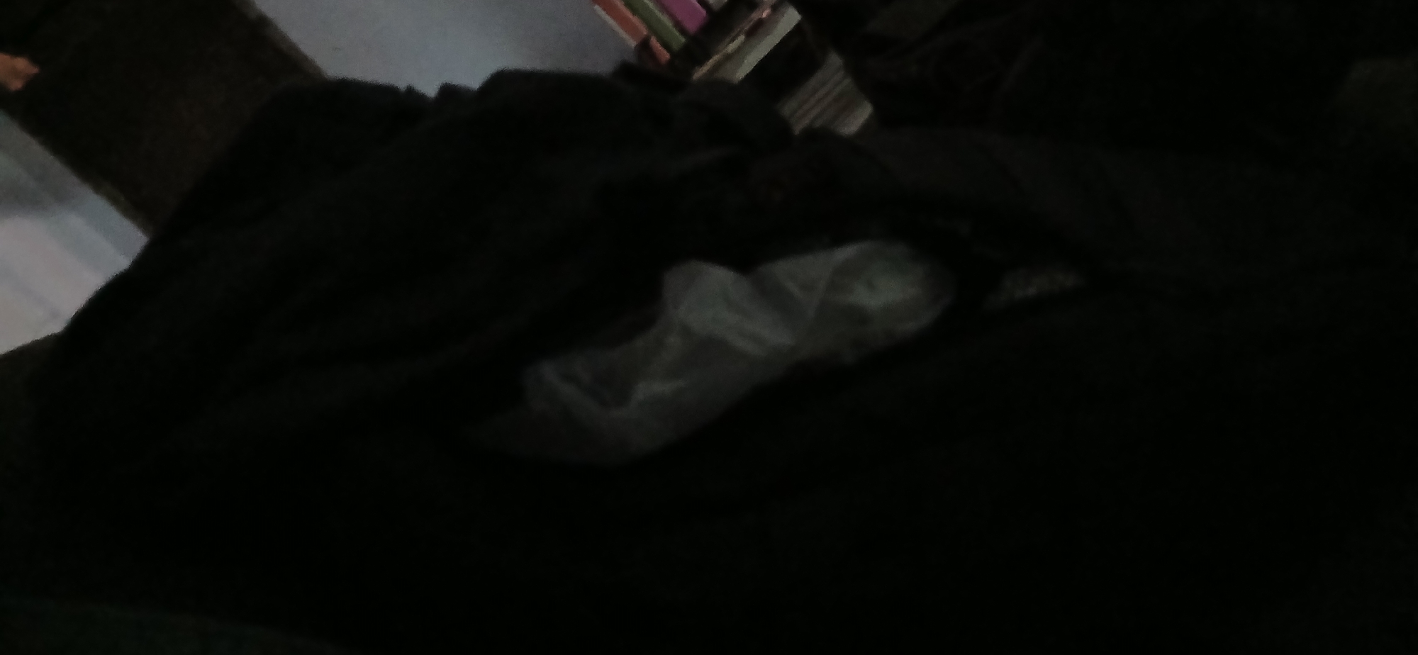 The image is dark and blurry, making it difficult to discern many details. It appears to be an indoor scene. In the foreground, there is a dark, textured surface, possibly fabric or clothing. To the right of this, a lighter, somewhat translucent object is visible, which might be a plastic bag. Further to the right and in the background, there are hints of shelves or stacked items, with some appearing to be books or boxes. The lighting is very dim, and much of the image is obscured by shadow. There are no discernible people or specific actions taking place. Due to the low quality and lack of clear features, it is not possible to determine the exact location, including its presence in Tenggarang, Indonesia.