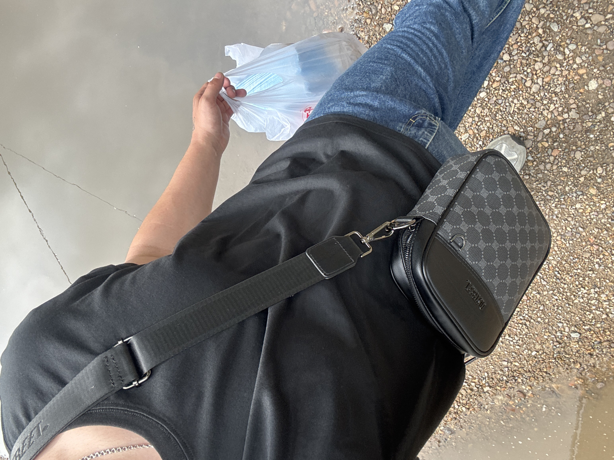 An individual, dressed in a black t-shirt and blue jeans, stands on a gravel surface adjacent to a shallow pool of water, possibly reflecting a cloudy sky. The person is carrying a plastic bag in their right hand and wearing a crossbody bag over their left shoulder. The crossbody bag is black with a patterned design and a silver clasp. The environment appears to be outdoors, with the ground composed of small pebbles and some areas of mud or standing water. The scene does not contain any specific visual cues that would allow for identification of the location as Birobidzhan, Russia.