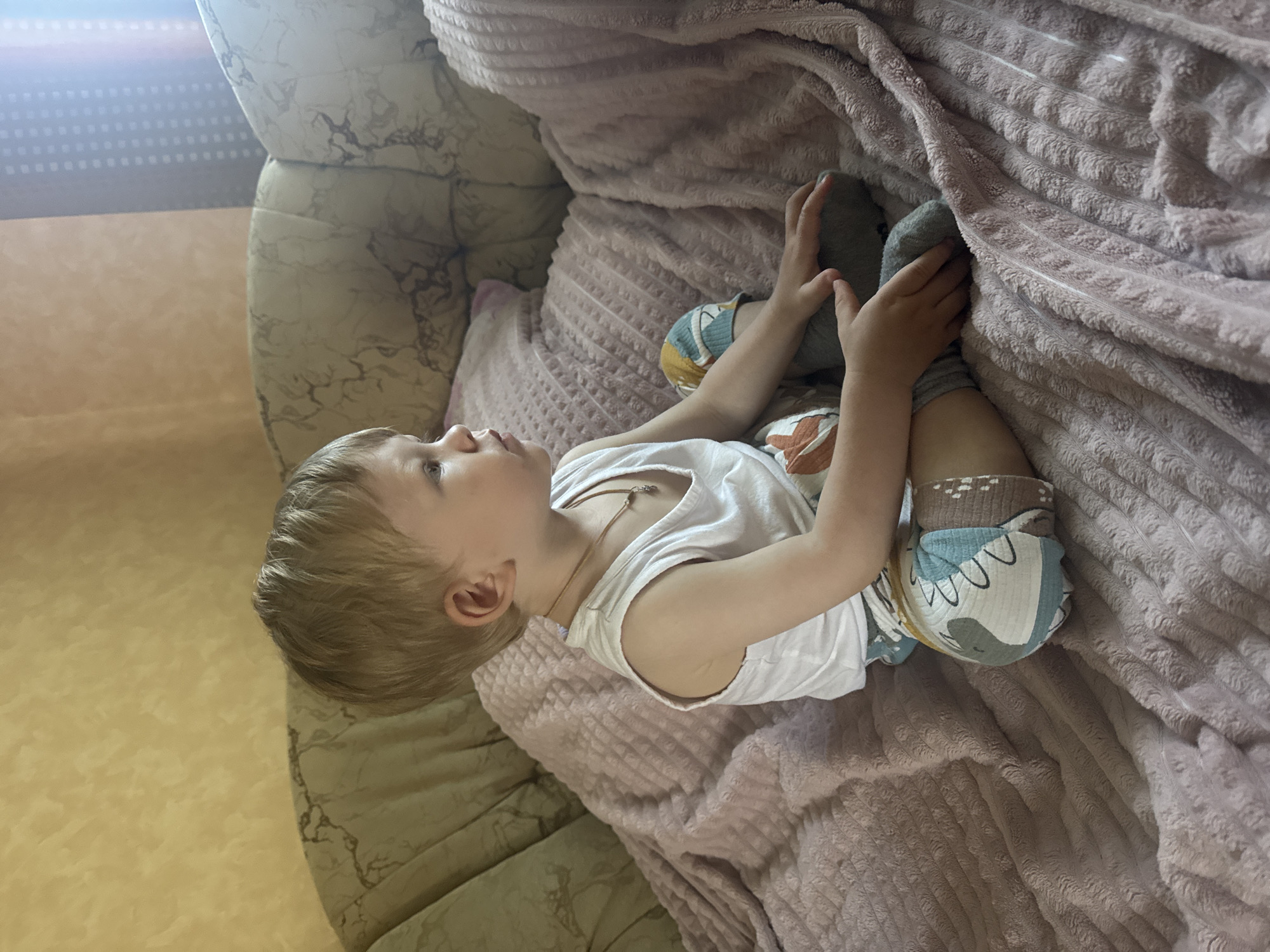 A young boy with blonde hair is sitting on a couch covered with pink blankets. He is wearing a white tank top and patterned shorts. The boy is looking to the right, with his hands resting on his legs. The couch appears to be in a living room with yellow textured walls and a window with blinds in the background. The overall scene suggests a casual indoor setting.