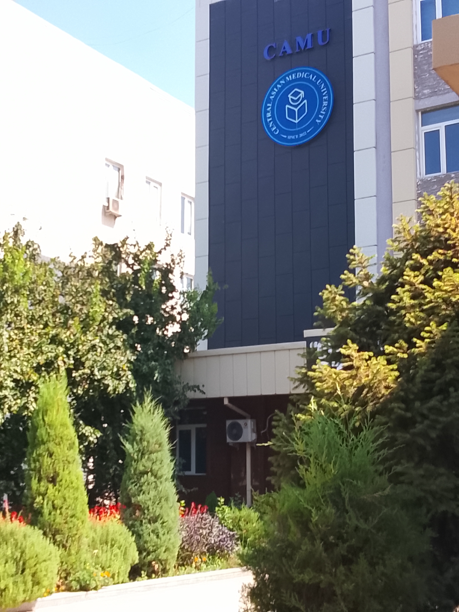 The image captures the facade of the Central Asian Medical University (CAMU) in Fergana City, Uzbekistan. The university building features a dark paneling section with the institution's name "CAMU" prominently displayed in blue letters at the top. Below the name, a circular blue logo is affixed, depicting an open book with an abstract representation of knowledge or learning. The text around the logo reads "CENTRAL ASIAN MEDICAL UNIVERSITY" and "SINCE 2019". The building has a modern architectural style with large windows visible on adjacent sections of the facade. In the foreground, lush green trees and manicured bushes with some flowering plants add a natural element to the scene. The overall impression is that of an educational institution set within a well-maintained environment.
