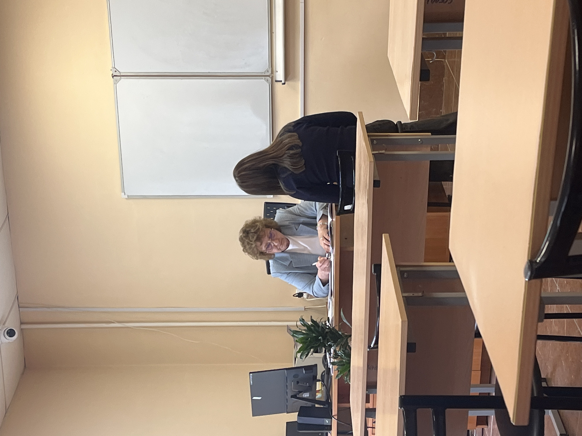 The image shows an interior scene, likely a classroom or office in Saint Petersburg, Russia, characterized by rows of desks and chairs. A woman with curly hair and glasses is seated at one of the desks, wearing a light blue blazer over a white shirt. She appears to be writing or working on something in front of her. Behind her, another individual with long brown hair is seated. A whiteboard is mounted on the wall behind them. A computer monitor, possibly a desktop, and a potted plant are visible on a desk further down the row. A security camera is noticeable on the wall near the ceiling. The scene suggests a setting of education or work.