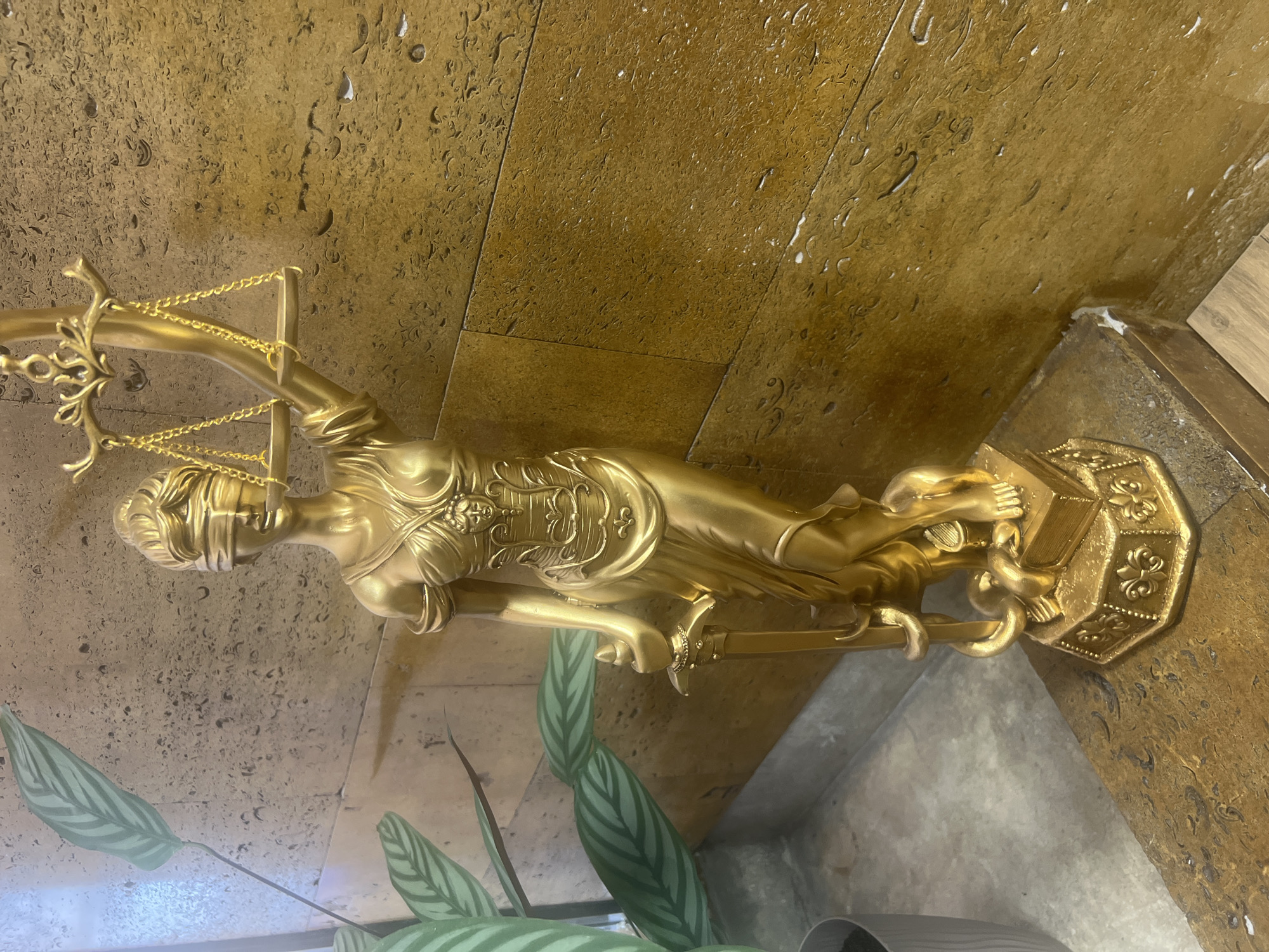 The media file features a golden statue of Lady Justice, positioned prominently in the center of the frame. The statue is blindfolded, holding a set of scales in her left hand and a sword in her right, which points downwards. Her attire includes decorative elements on the chest, and she stands on an ornate, octagonal base with embossed patterns, resembling fleur-de-lis.
The background comprises a textured, golden-brown surface, likely a wall or paneling, exhibiting an irregular, pockmarked, or fossil-like pattern, with visible seams suggesting it is composed of tiles or large panels. The floor or a lower surface visible towards the bottom and right is a lighter, grayish-brown tone with some reflectivity.
In the foreground, to the left of the statue, are several large, patterned green leaves belonging to a plant, possibly a Calathea, with distinct striped markings. A portion of a dark plant pot is visible at the very bottom left corner.
The scene is static, depicting no activity or event. The lighting suggests an indoor setting, and no specific time of day can be determined. There is no visible text in the image. The overall impression is one of formal decoration, possibly in an institutional or commercial environment.