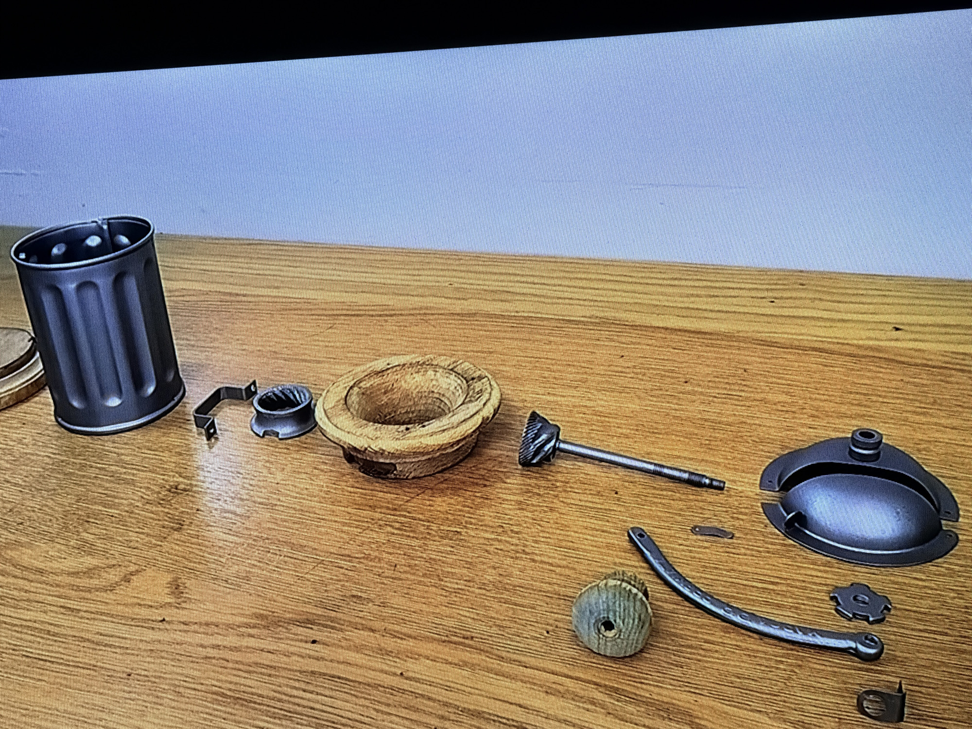 An assortment of disassembled parts from a coffee grinder are laid out on a wooden table. The parts include a dark grey cylindrical hopper with vertical grooves, a metallic clamp, a wooden bowl-shaped component, a metal shaft with a grinding burr, a small flat metal piece, a wooden knob, a curved metal handle with perforations, a small gear-like metal piece, and a dark grey domed lid with a handle. The background consists of a plain light blue wall. The scene is located in Castillo Bajo-conejito, Spain.