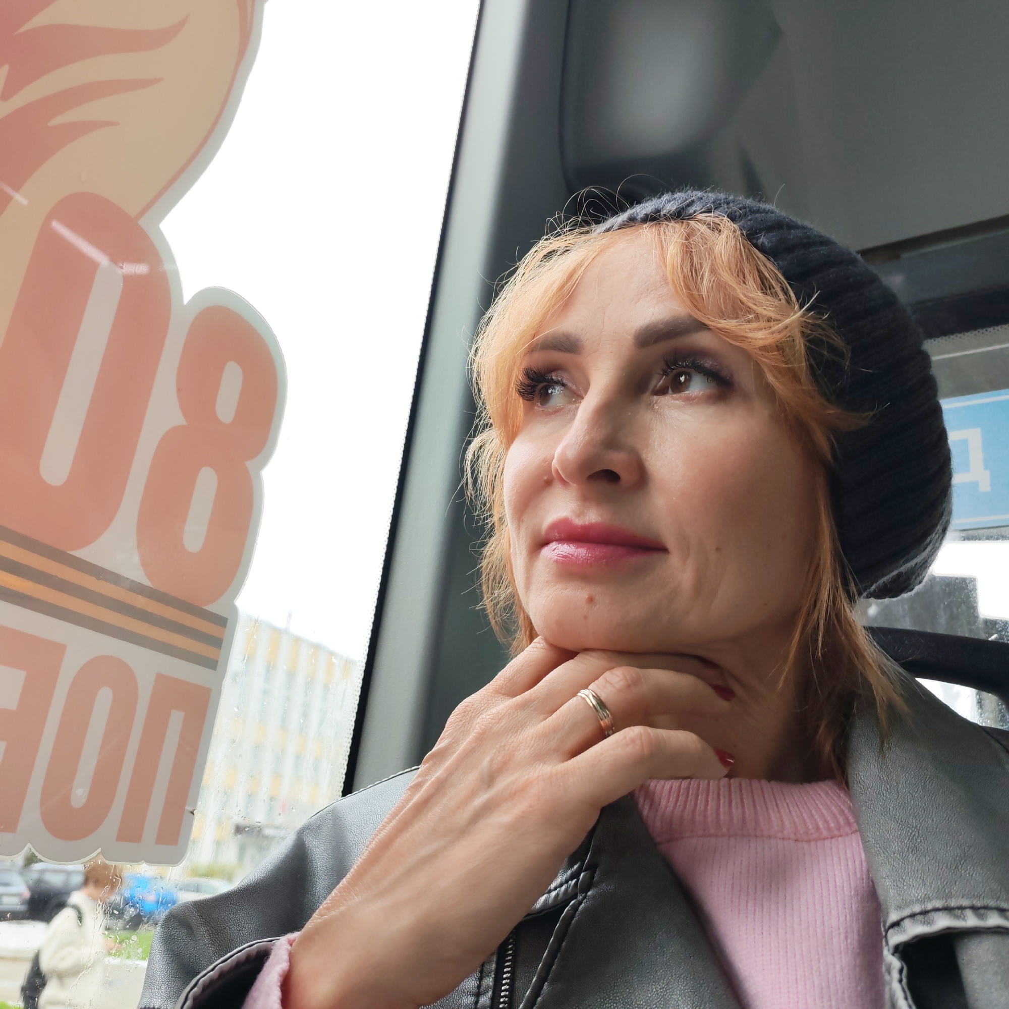 A middle-aged woman with reddish-blonde hair and a dark knit hat is seated inside a vehicle, likely a bus or train, looking out the window. She is wearing a pink sweater and a dark leather jacket. Her right hand is raised to her neck, and a wedding ring is visible on her finger.
Through the window, a blurry view of a street is visible, and a large sticker with pink and white letters, which appears to be part of an advertisement, is partially obscuring the view. The sticker also has a geometric pattern in brown and white. Based on the "ПОП" and the sticker commemorating victory in the Great Patriotic War (WWII), and the architectural style visible through the window, it is highly likely this photo was taken in a Russian city. Using reverse image searches of the sticker, it is possible that this image was taken in the city of Chaikovsky, Russia.