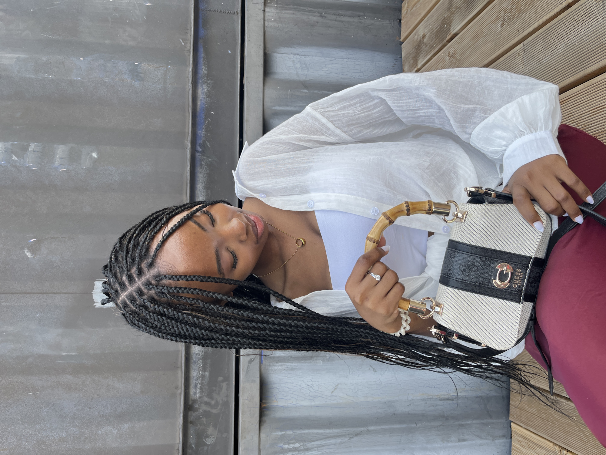 An outdoor portrait features a young Black woman with long, braided hair, wearing a white tube top and an unbuttoned white linen shirt. She is holding a beige and black Guess handbag with a bamboo handle. She is seated on a wooden bench in front of a corrugated metal wall. Her left hand, adorned with a ring and bracelet, is touching the handbag's handle. The scene suggests a casual, urban setting. There is no specific information to suggest the location is Kazan, Russia.