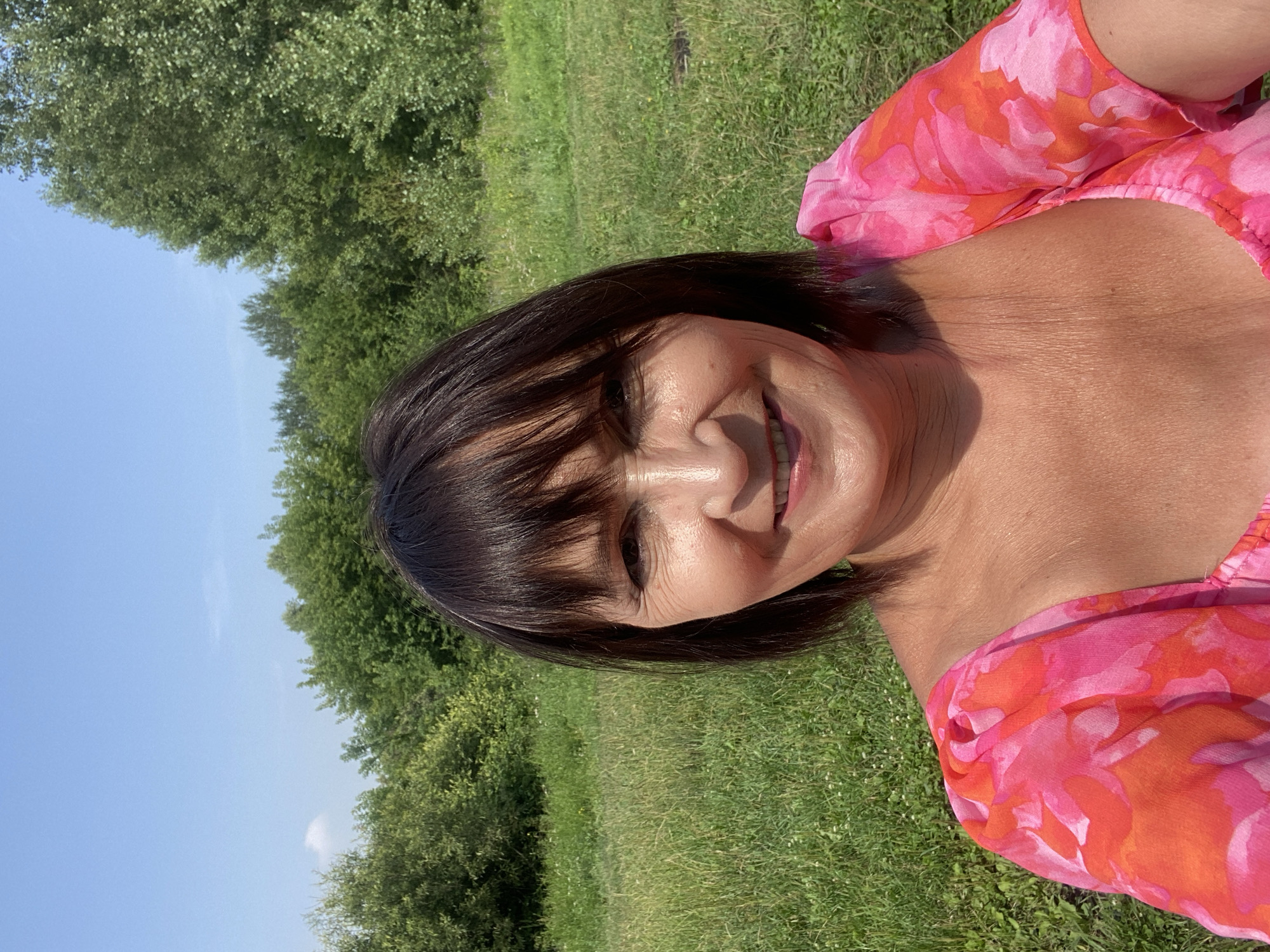 A woman with dark hair and bangs, wearing a pink and orange floral dress, smiles at the camera in a selfie. She is outdoors, with a backdrop of green grass and trees under a clear blue sky. The location is Novokuznetsk, Russia.