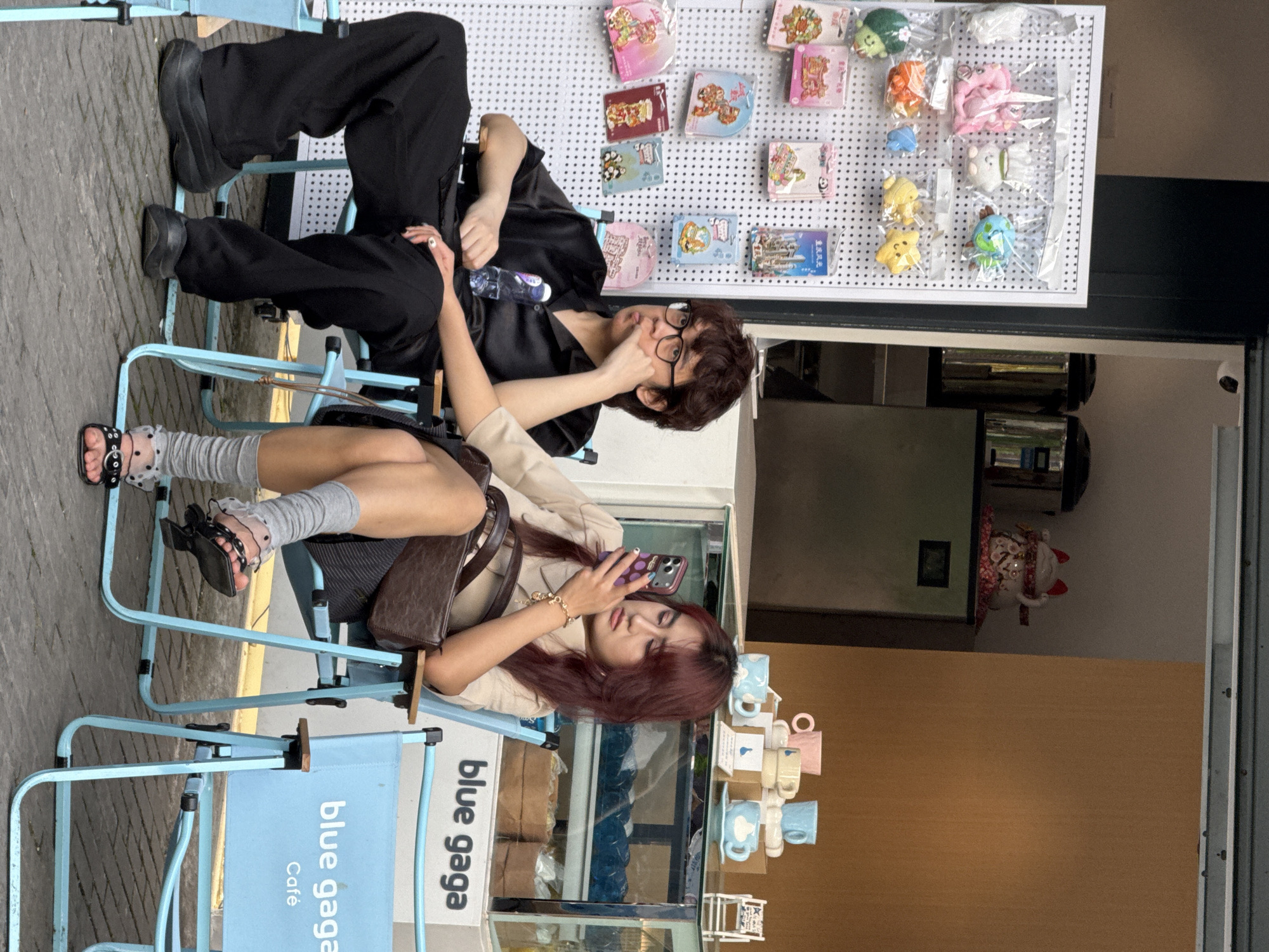 Two people are sitting in blue folding chairs in front of a building displaying a "blue gaga Café" sign. The person in the chair further from the building is wearing black pants, a black top, and black shoes. They are also wearing glasses and have brown hair. They have one hand on their forehead. The person in the foreground chair is wearing a tan jacket, a grey and black skirt, grey socks, and black sandals. They have a brown handbag and are looking at their phone. A white pegboard with various small items is mounted on the building behind the people. The scene is located in Yuzhong District, China.
