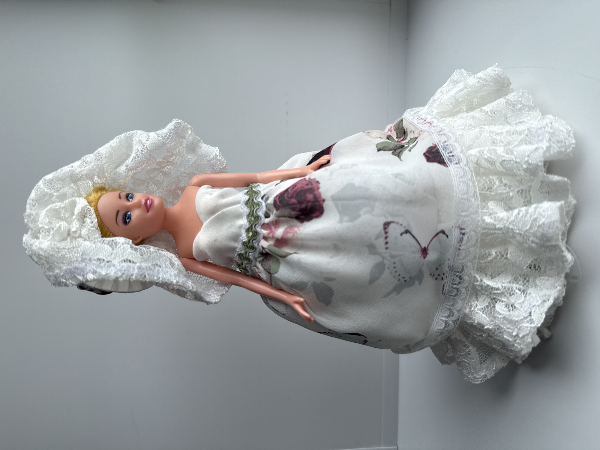 A Barbie doll lies on its side in what appears to be a light grey or white room, possibly a shelf or a display case. The doll has blonde hair and is wearing a white strapless dress with a floral print consisting of burgundy and green roses. The dress is trimmed with white lace at the top and bottom. The doll is also wearing a large white lace headpiece or hat. No city name or location can be definitively determined from the image.