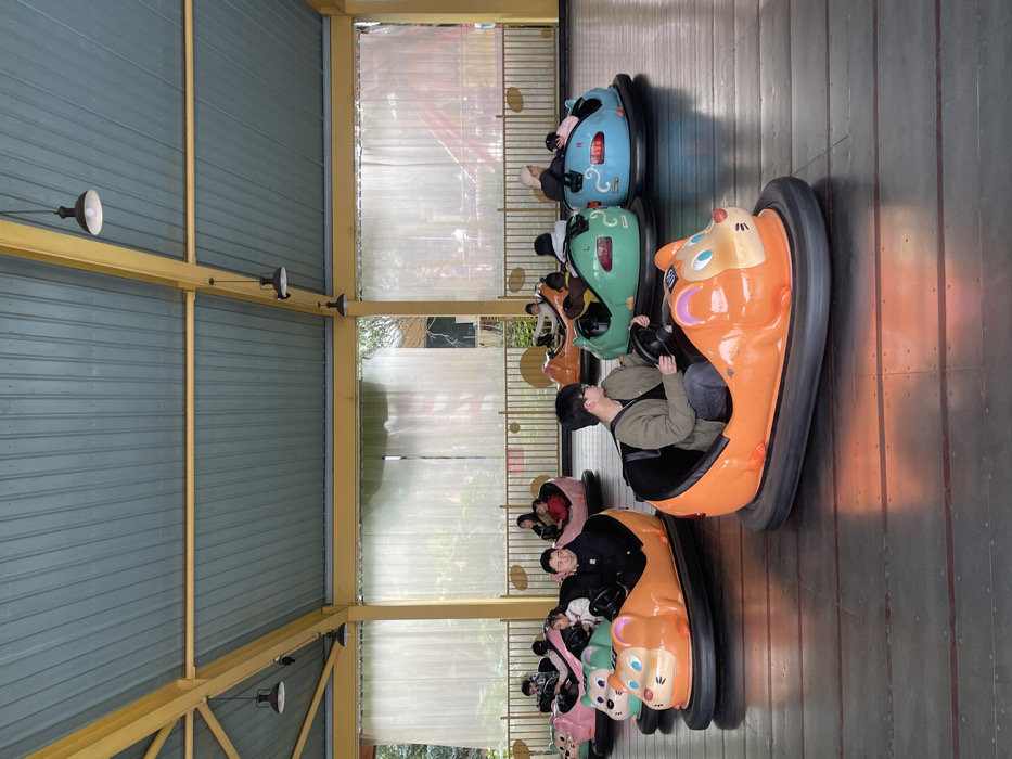 The image displays an indoor bumper car arena located within the Chongqing High-tech Zone, China. The structure features a blue-grey corrugated metal roof supported by yellow structural beams, from which multiple black industrial-style pendant lights are suspended. The arena's perimeter is enclosed by translucent corrugated barriers, revealing a green, treed exterior and distant architectural elements. The floor is a dark, reflective surface.
Approximately 10-12 individuals, consisting of adults and children, are observed seated in various animal-themed bumper cars, predominantly designed as cartoon mice or rats. The cars are colored orange, pink, green, and light blue, each equipped with a black rubber bumper surrounding its base. In the immediate foreground, a young male wearing glasses and a light green jacket is seated in an orange bumper car, grasping the steering wheel and looking to his left. Adjacent to him, another orange bumper car contains an adult male in a black jacket and a child passenger. Other cars are scattered across the arena, occupied by individuals or pairs holding steering wheels, indicating active participation or preparation for the bumper car ride. No active collisions are depicted.