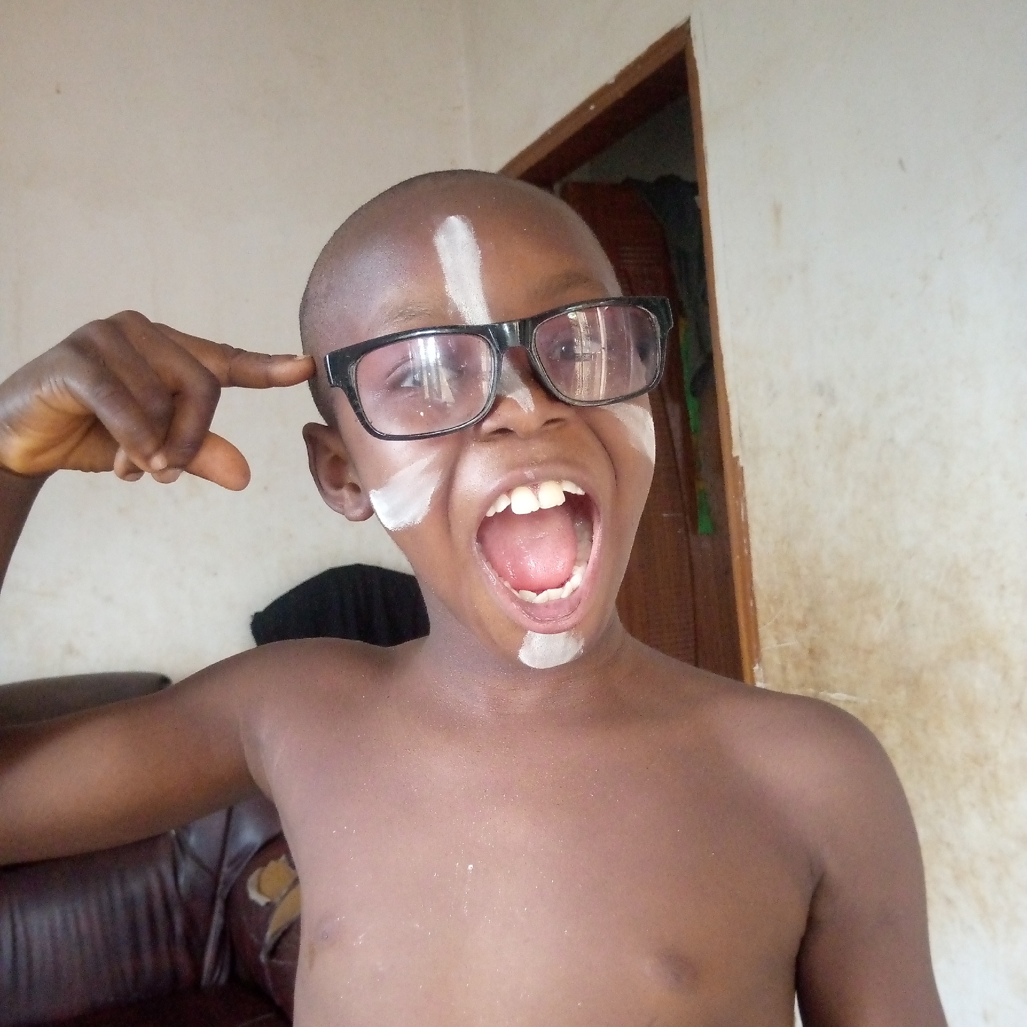 A medium shot captures a young, bald child with dark skin posing energetically indoors. The child wears black-framed glasses and has distinctive white paint or makeup applied to his face, including a vertical stripe on his forehead, a stripe on each cheekbone, and a small mark below his lower lip. He is shirtless, revealing his bare chest and shoulders, with a small white smudge visible below his neck.
The child's left hand is raised, with his index finger pointing to his right temple in a gesture often associated with thinking or a "mind-blown" expression. His mouth is wide open in a loud yell or shout, showcasing his tongue and both upper and lower teeth, conveying an emotion of excitement, playfulness, or even a staged scream.
The background features a plain, light-colored wall, possibly beige or off-white, which exhibits some minor stains or wear. To the right of the child, a dark brown wooden door frame is visible, leading to a darker interior space. To the lower left, a section of a dark brown, possibly worn leather sofa or armchair is partially in view. The scene is lit by what appears to be natural daylight. No text is visible in the image.