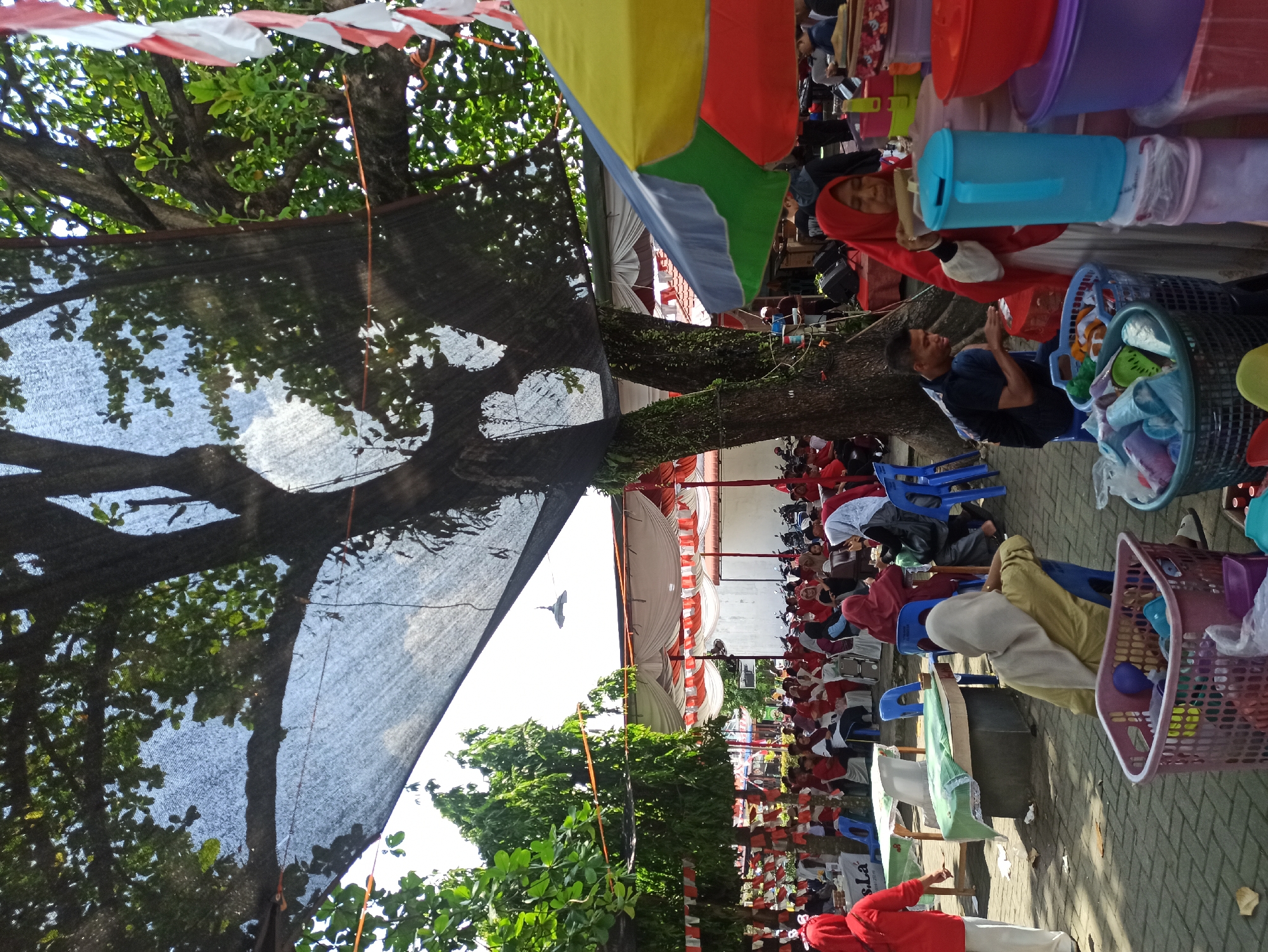 An outdoor scene is depicted with many people seated under a large tree shaded by a dark mesh covering, suggesting a public gathering or event. Red and white Indonesian flags are strung along the edge of a tent structure in the background, where more people are seated on chairs. Several tables are visible, some draped with white cloths and one with a green cloth. In the foreground, a woman wearing a red hijab is interacting with a stack of containers, and another person wearing a yellow hijab is seated on a blue chair. Baskets filled with plastic items are on the ground. The ground is paved with interlocking bricks, and more trees and foliage are present. The atmosphere appears to be that of a community event, possibly a festival or a market, given the presence of vendors and numerous attendees.