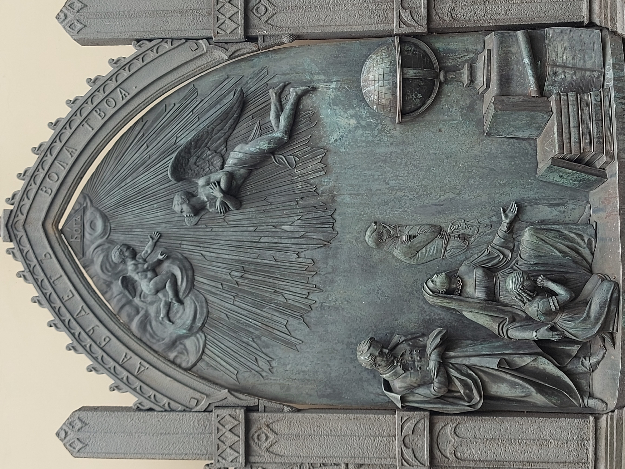 The image shows a close-up of a bronze relief sculpture, part of a larger architectural feature. The relief depicts several figures in a scene suggestive of death and ascension. On the left side, a cherubic figure rises from clouds with radiating lines. Above, an angel with wings descends, arms folded. To the right, three figures recline in what appears to be a death scene. A woman holds a child. A stack of books and a globe are also present. The inscription "Bora Tboa" can be read on the arch surrounding the left side of the relief. This relief is likely located in Saint Petersburg, Russia, as it appears on one of the doors of the Chapel of the Ascension.
