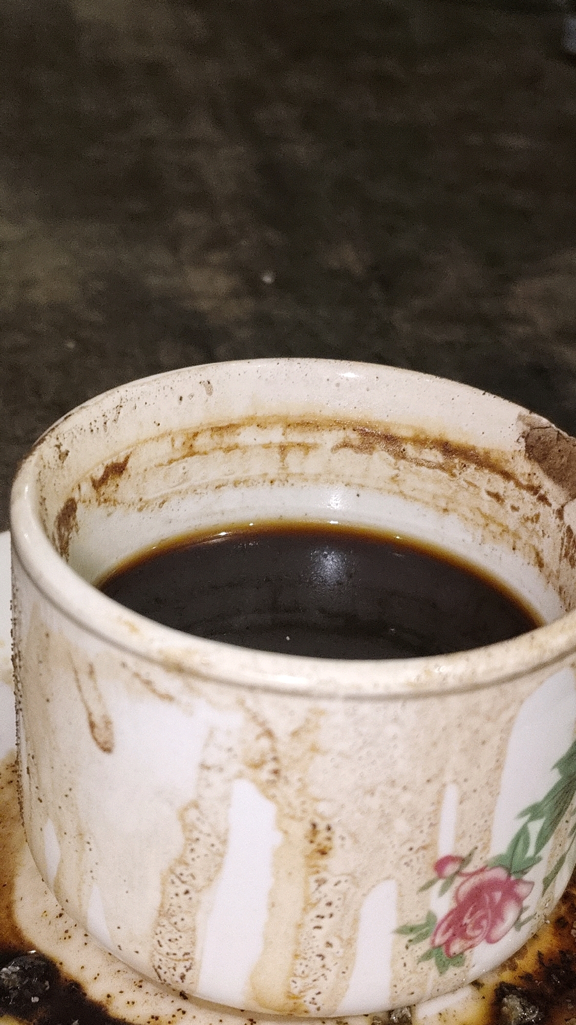 The image displays a close-up, high-angle shot of a partially consumed cup of dark coffee. The cup itself is white with a floral design, featuring a pink rose and green leaves. The rim of the cup and its exterior are stained with dried coffee residue, creating streaks and patches of brown. The coffee within the cup appears dark and opaque. The cup sits on a saucer, which also shows signs of coffee spillage. The background is out of focus, showing a dark, textured surface, likely a table or countertop, with a muted, earthy tone. The lighting appears to be dim, casting a slight shadow. The setting is a domestic environment in Karangtalun, Indonesia.