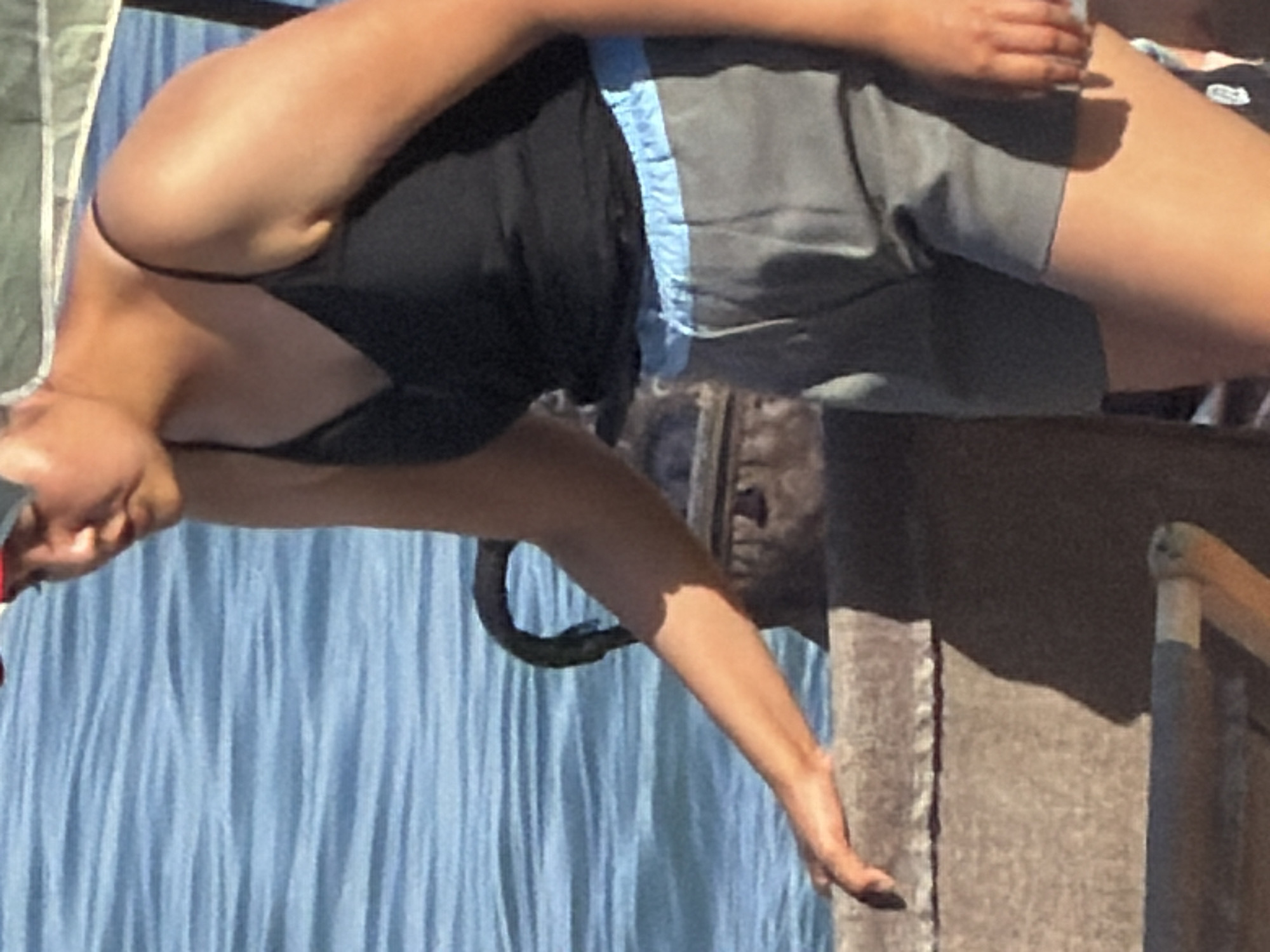 A medium shot shows a person in a black camisole and gray shorts with a blue drawstring standing on a concrete ledge near a body of water. The person is leaning on the ledge with their left arm and hand. The person is wearing a red hat that is not fully visible. The water is blue and appears to be rippling. A dark-colored object, possibly a bag, is behind the person. The scene is outdoors and appears to be during the day.