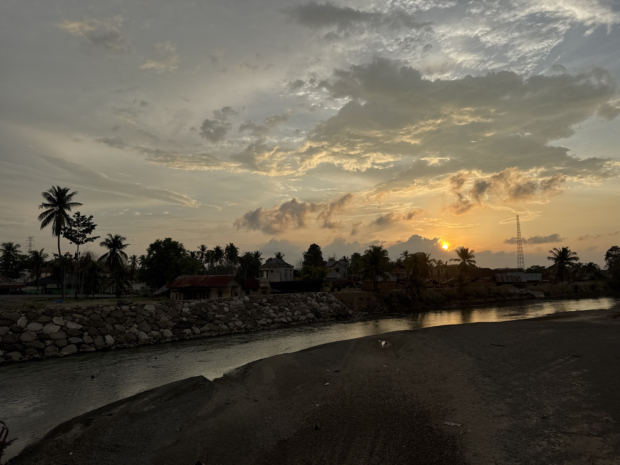 An outdoor scene at sunset depicts a tranquil riverside in the city of Meunasah Pupu, Indonesia. The sky is a canvas of warm hues, transitioning from soft oranges near the horizon to muted grays and blues above, punctuated by dramatic, textured clouds. The setting sun, a bright orb partially obscured by clouds, casts a golden glow on the scene, reflecting on the surface of the water.
The river flows from left to right across the image, its surface rippled by a gentle current. The left bank of the river is reinforced with a sturdy retaining wall constructed from large, irregular stones. Beyond this wall, a cluster of buildings, likely houses, are visible amongst lush green palm trees. These structures have varied roofing, some appearing to be red tile and others darker materials. A tall communication tower stands to the right in the background, silhouetted against the vibrant sky.
The foreground is a dark, sandy embankment that slopes down towards the river. The sand is coarse and appears slightly damp in places. Scattered debris, such as small twigs and pieces of paper, are visible on the embankment. The overall atmosphere is one of peaceful evening serenity, characteristic of a coastal or riverside settlement in Indonesia. No people are visible in the frame.