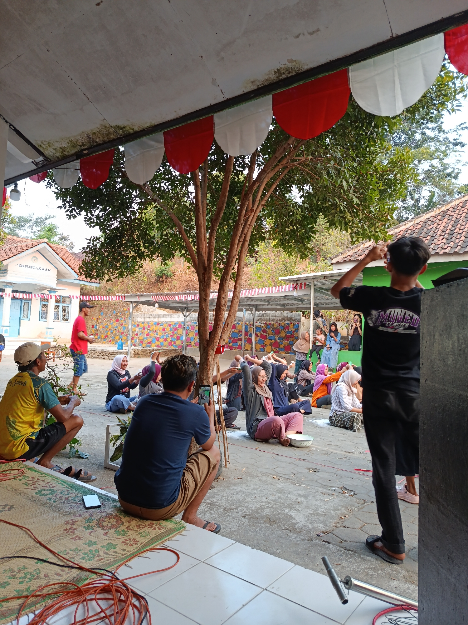 The image depicts an outdoor scene in Pocol, Indonesia, likely a community gathering or event. In the foreground, a group of people are seated on the ground, many appearing to be participating in an activity, with some raising their arms. One individual in the center is laughing. A man in a yellow shirt and a man in a blue shirt are also visible in the foreground, observing the activity. To the right, a young man in a black t-shirt is standing and appears to be taking a photo with his phone. In the background, a building with "PERPUSTAKAAN" (Library) written on it is visible, suggesting the event is taking place near or at a public library. Red and white banners are hanging overhead, potentially indicating a national celebration or holiday. The environment appears to be paved with a tree providing shade. The overall atmosphere seems to be one of community engagement and enjoyment.
