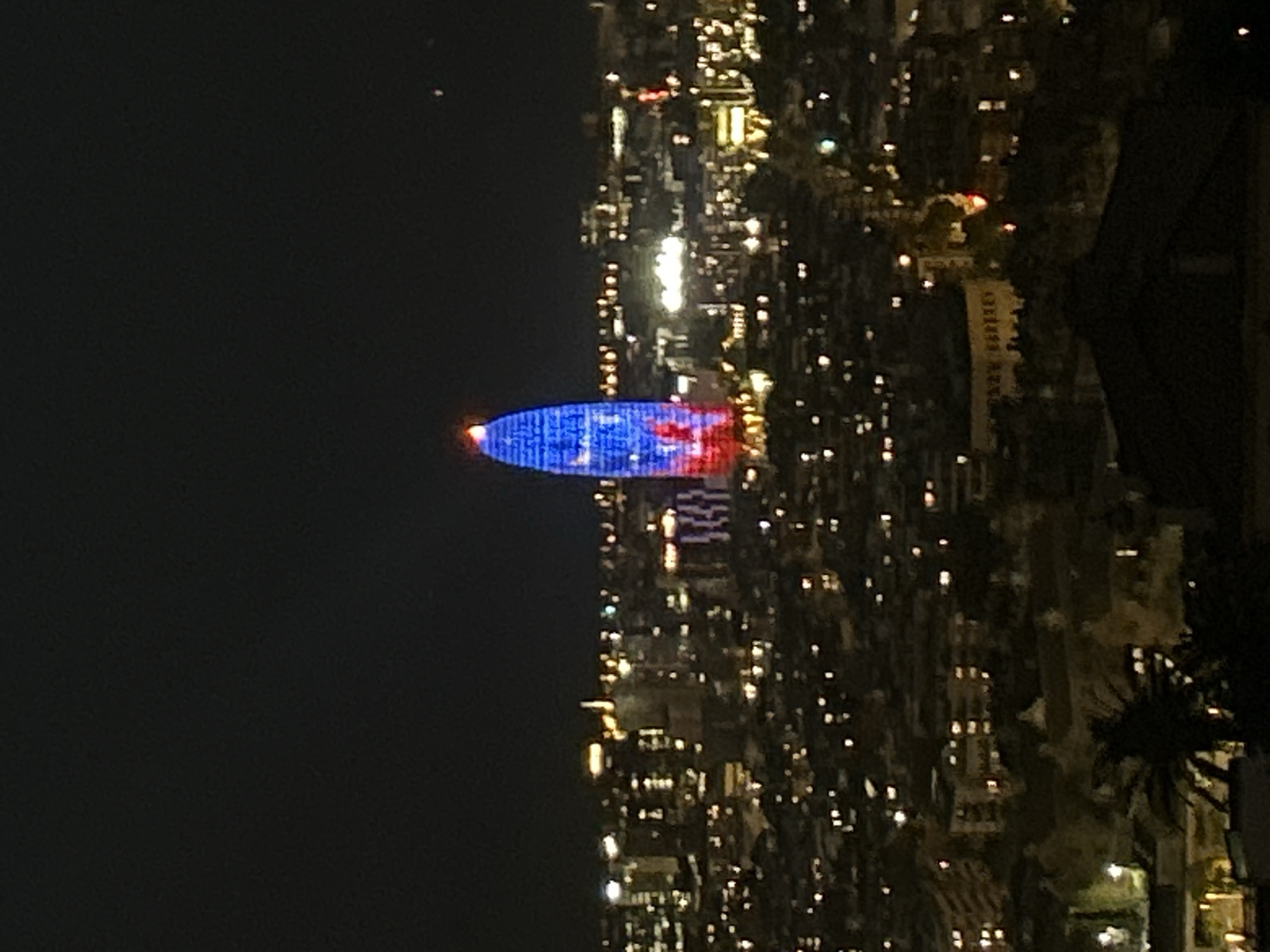 The image shows a night view of Barcelona, Spain, focusing on the Torre Glòries illuminated in blue and red. The tower's shape is distinct, rising above the cityscape. The surrounding area is lit by numerous city lights, creating a sparkling effect across the urban landscape. The sky is dark and clear with one visible light point. Palm trees and buildings can be seen towards the right of the frame.