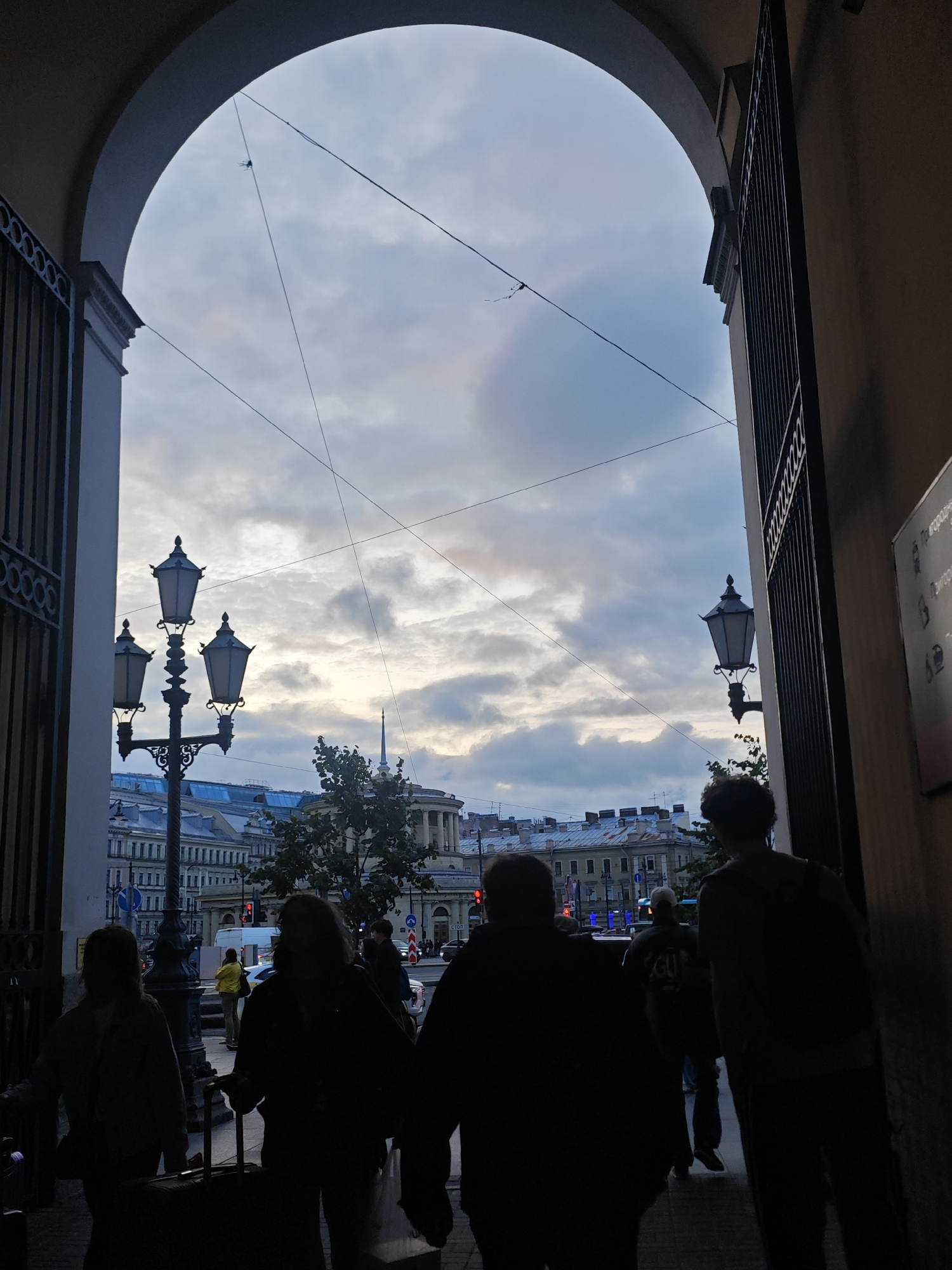 This is a vertical, medium shot photograph taken from within an archway in Saint Petersburg, Russia. Silhouetted figures are walking through the archway, some with luggage. The sky above is cloudy with hues of blue, grey, and pink. In the distance, classic Saint Petersburg architecture is visible, including ornate buildings and a spire. Two street lamps, characteristic of the city, are illuminated on either side of the archway. Wires crisscross the sky, a common sight in the city. The scene captures a moment of movement and urban life under a dramatic sky.