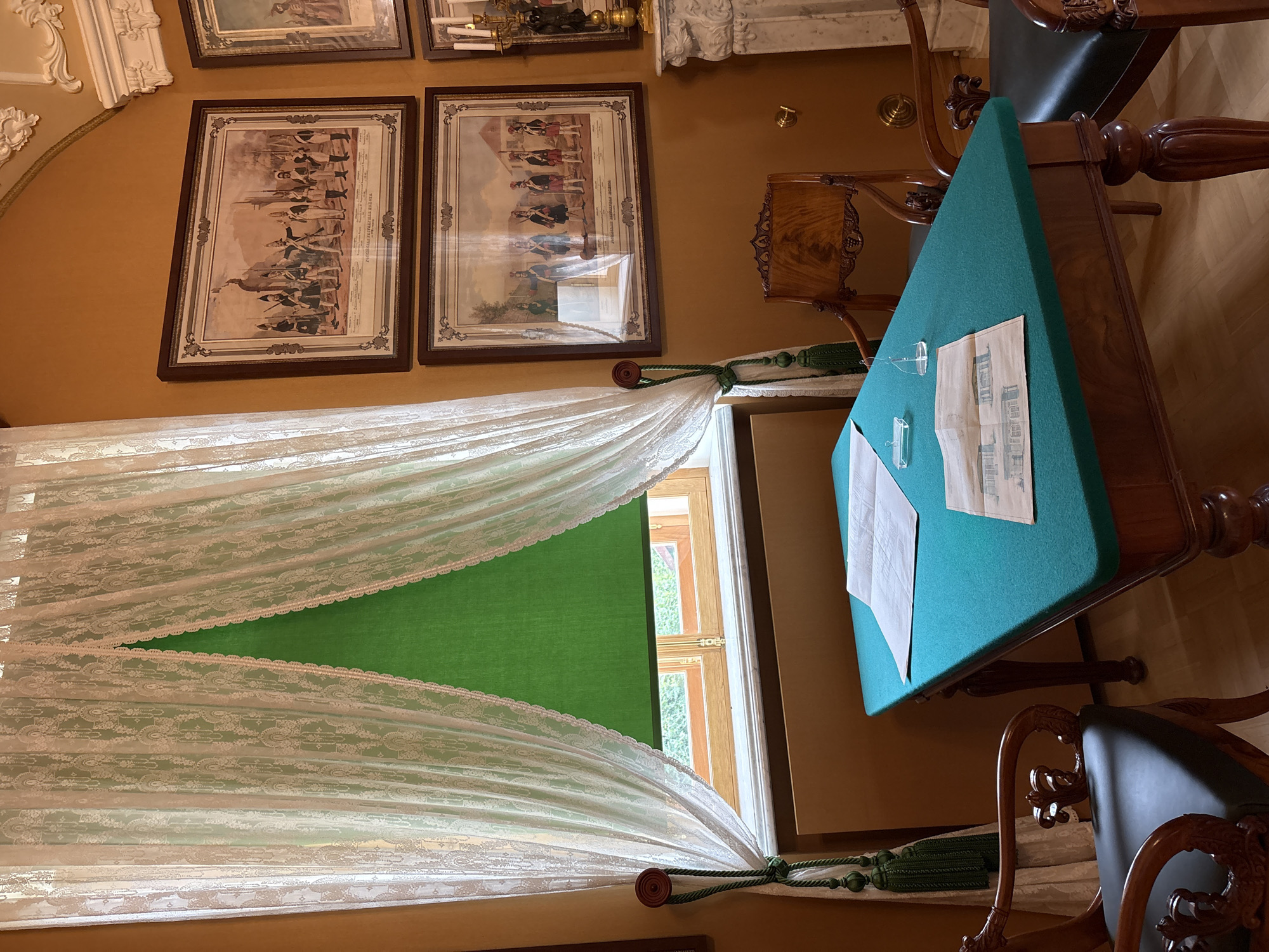 The image depicts an interior scene, likely a historical room within the Gatchina Palace in Gatchina, Russia. The room features ornate decor with a warm, ochre-colored wall. A large window is covered by sheer white lace curtains with decorative green tassels and a bright green roller blind. On the window sill, a beige radiator cover is visible.
In the foreground, a dark wooden table with a rich teal felt top is set. On the table are two pieces of paper, one appearing to be a floor plan or architectural drawing, and another smaller document. A small clear glass object, possibly a paperweight or a small vase, sits on the table as well.
Two antique wooden chairs with dark upholstered seats are positioned around the table. One chair, partially visible on the left, has elaborately carved arms and a high, decorative back. Another chair, to the right, is also intricately carved. In the background, another ornate wooden chair is placed.
The walls are adorned with three framed prints, depicting historical military figures in uniform. A decorative marble fireplace with a gilded candelabra and a statue is visible on the right side of the image. The flooring appears to be polished wood. There are no people present in the image.
