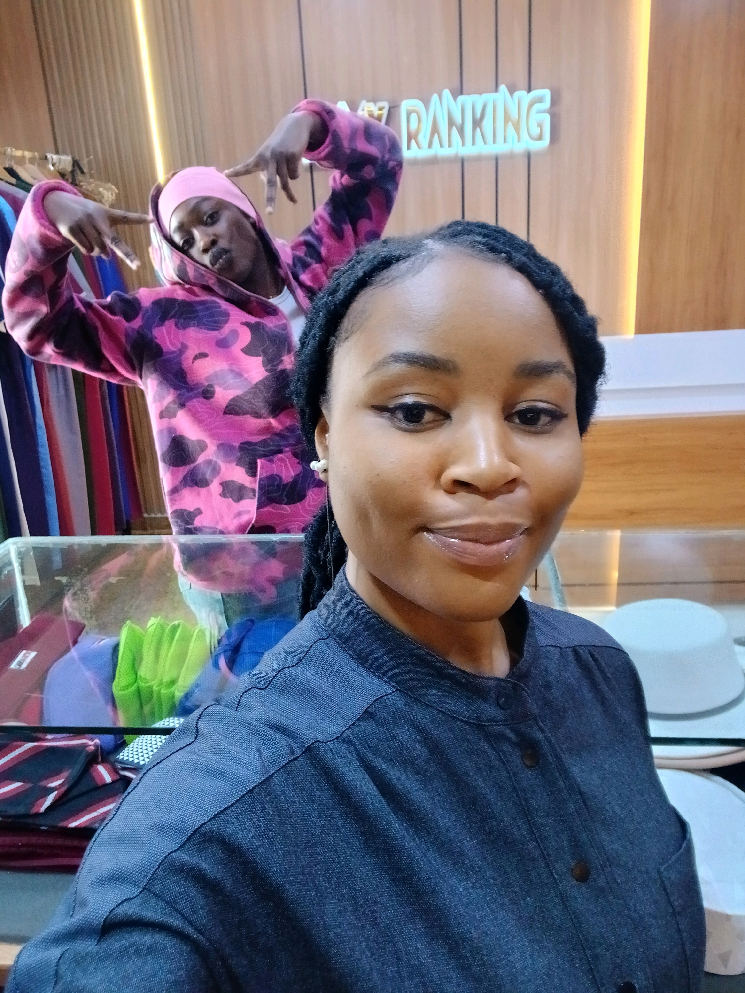 Two individuals are present in an indoor retail environment. The foreground features a woman facing the camera, her head and shoulders visible. She has dark skin, dark braided hair, and is wearing a dark-colored collared shirt with buttons. She has on eyeliner and a pearl earring. In the background, a second person, appearing to be of darker complexion, is wearing a pink and purple camouflage hoodie and a pink head covering. This person is striking a pose with their hands. Behind them, a sign with illuminated lettering that reads "RANKING" is mounted on a wooden wall. The background also shows clothing racks with garments and a glass display case containing various items. The lighting appears to be artificial, with a prominent warm light source behind the "RANKING" sign. The overall scene suggests a retail store setting.