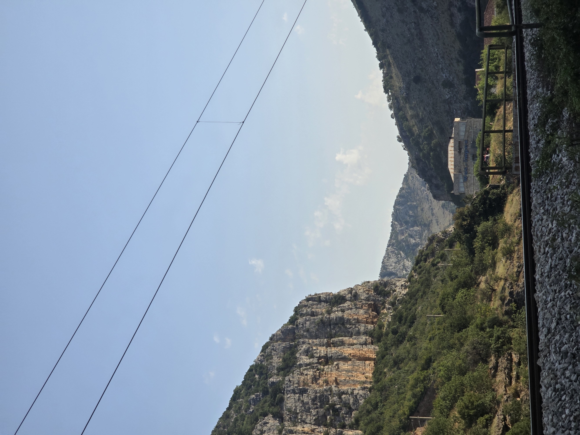 The image depicts a mountainous landscape under a clear blue sky with a few wispy clouds. Two parallel dark power lines stretch diagonally across the upper portion of the frame. In the mid-ground, a building with a distinctive glass facade and a tiled roof is nestled amongst the hills. In the foreground, a section of railroad track with gravel ballast runs horizontally across the lower part of the image. The surrounding environment is characterized by steep, rocky inclines covered in dense green vegetation, with some exposed rock faces showing shades of orange and brown. The scene appears to be an outdoor, natural setting, possibly a canyon or valley, with man-made elements like the power lines and railroad tracks indicating human presence and infrastructure.
