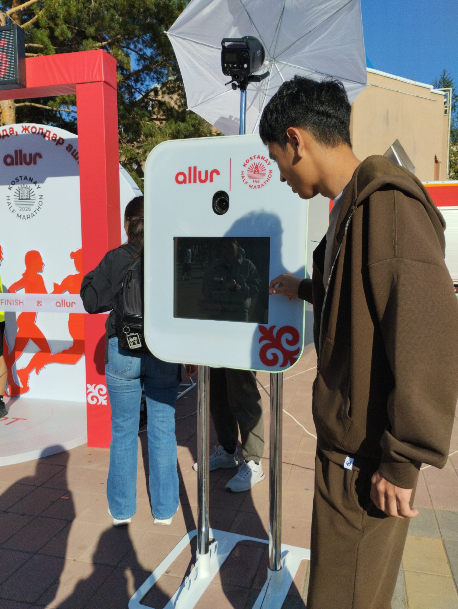 The image captures an outdoor scene at what appears to be a start or finish line for a half marathon in Kostanay, Kazakhstan. In the foreground, a young man in a brown hooded tracksuit is interacting with a white photo booth, his right hand extended towards the screen. The photo booth has the "allur" logo and the "KOSTANAY HALF MARATHON 2026" text. To the left, a red archway structure is visible, also branded with "allur" and featuring graphics of runners and the word "FINISH". Behind the man, a woman wearing a black jacket and jeans with a backpack is partially visible, facing away from the camera and towards the photo booth. In the background, a white umbrella-like diffuser is positioned above the photo booth, attached to a light stand with a camera. The environment appears to be paved with brick-like tiles, and there are shadows cast on the ground, indicating it's a sunny day. In the upper right corner, a building with light-colored walls and a tan roof is visible. The overall scene suggests a lively event, likely a sporting competition.