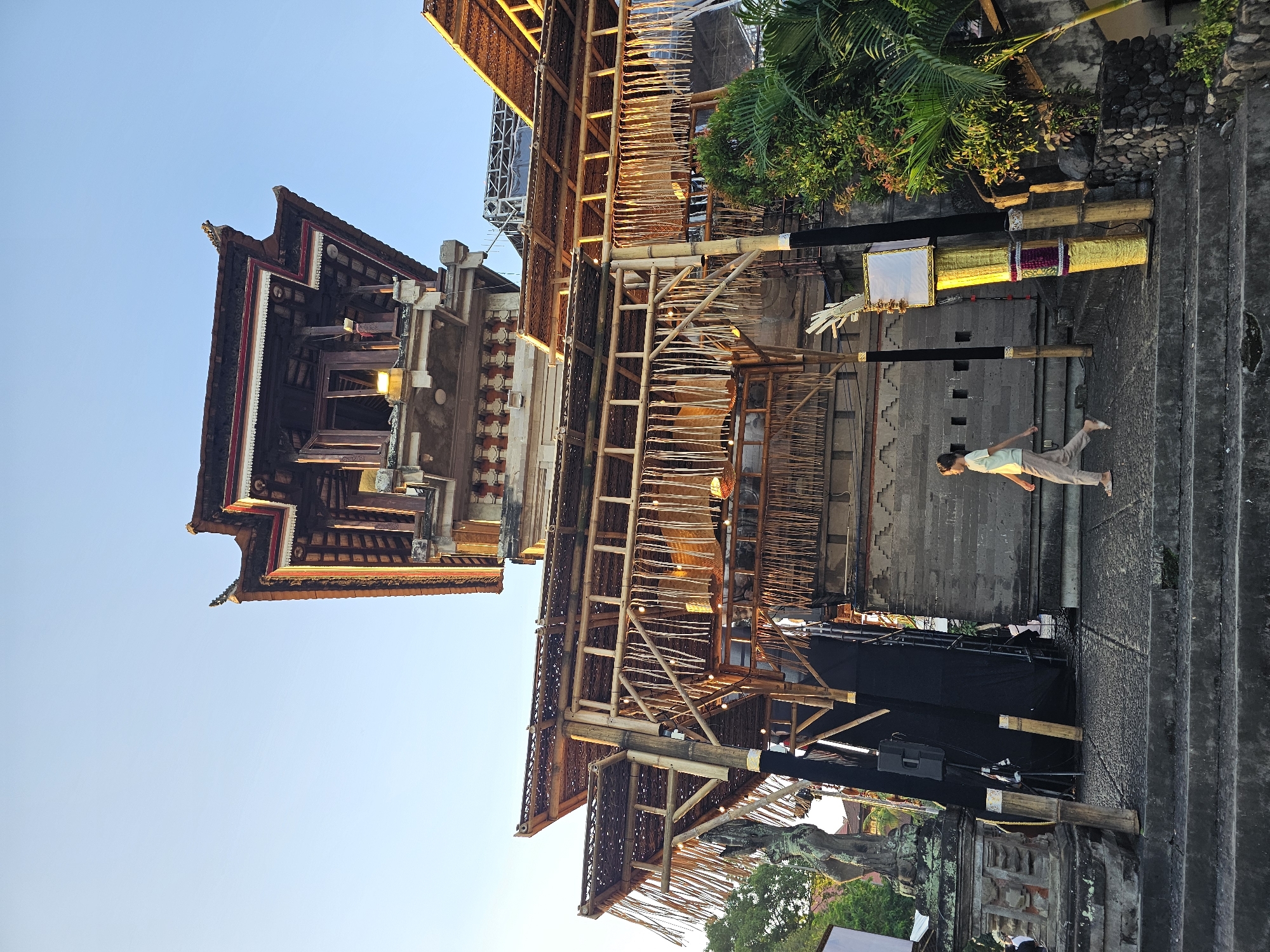 A low-angle, full shot captures a traditional Balinese structure against a clear, pale blue sky in Denpasar, Indonesia. The multi-tiered building features ornate wooden roofing with multiple layers and intricate carvings. Bamboo structures with thatched roofs, adorned with hanging, woven decorations and string lights, extend from the main building, creating an entranceway. A woman in a light-colored t-shirt and loose pants walks across stone steps towards the building. To the left of the steps, a weathered stone statue stands on a pedestal. In front of the entrance, two tall, bamboo poles are decorated with gold and purple accents, topped with a white rectangular sign. Lush green foliage, including palm trees, surrounds the right side of the structure. The scene appears to be set up for an event, with black fabric panels and stage lighting visible.