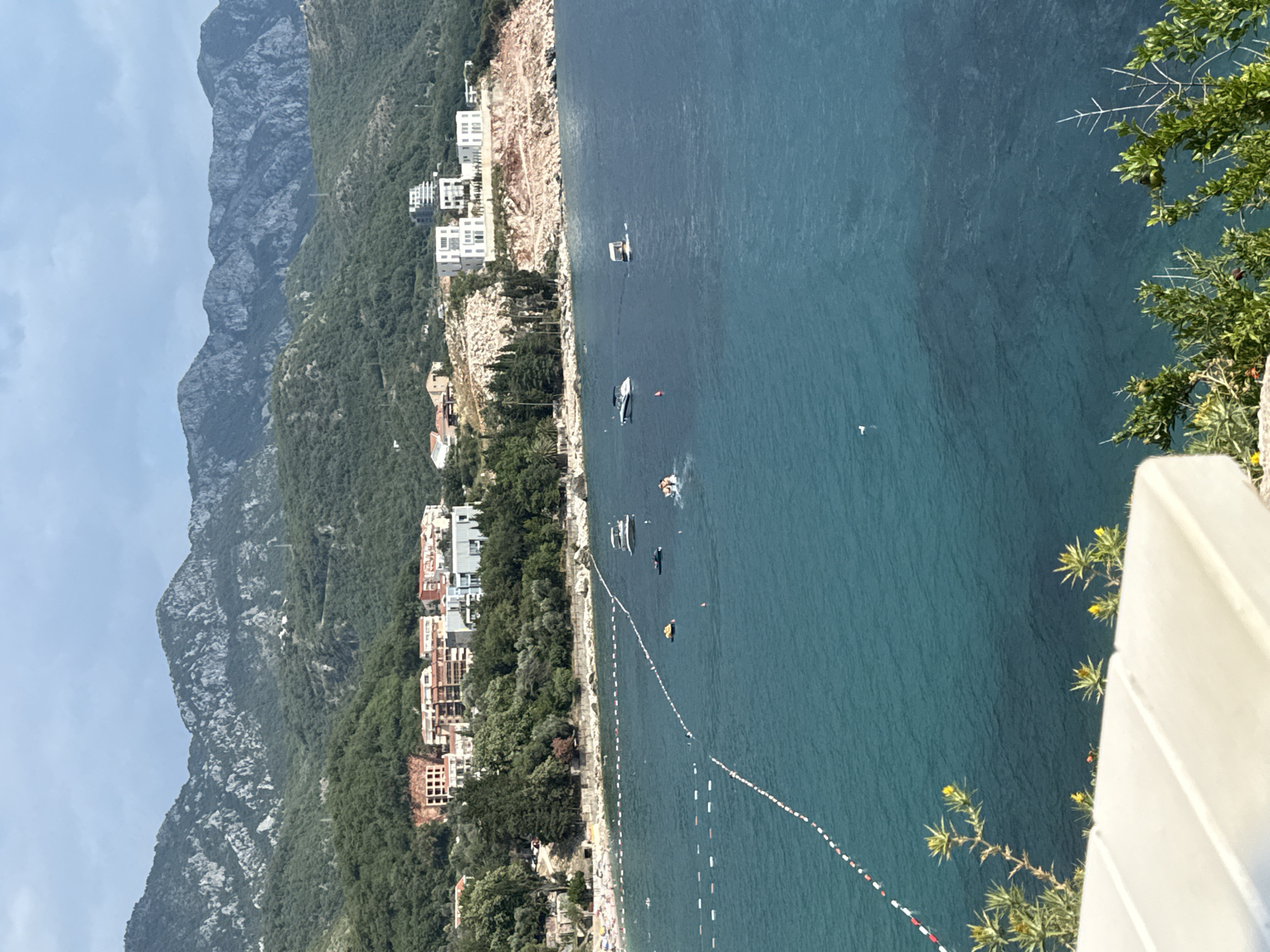 This media file captures a scenic view of Kamenovo, Montenegro. The scene features a coastline with a rocky shore, dense green vegetation, and several modern buildings nestled into the hillside. The turquoise water of the Adriatic Sea stretches out, with several boats dotted on its surface, suggesting recreational activity. A marked swimming area with buoys is visible in the water, indicating a popular beach. The image also shows a sliver of a beach with people enjoying the sun, hinting at a warm and pleasant day. The background is dominated by imposing, rugged mountains that rise steeply from the coast, their slopes covered in greenery. The sky is partly cloudy, suggesting it's likely daytime, possibly late morning or early afternoon, given the lighting. The overall atmosphere is tranquil and picturesque, highlighting the natural beauty of the location combined with the human presence of settlements and leisure activities. There is no visible text in the image.