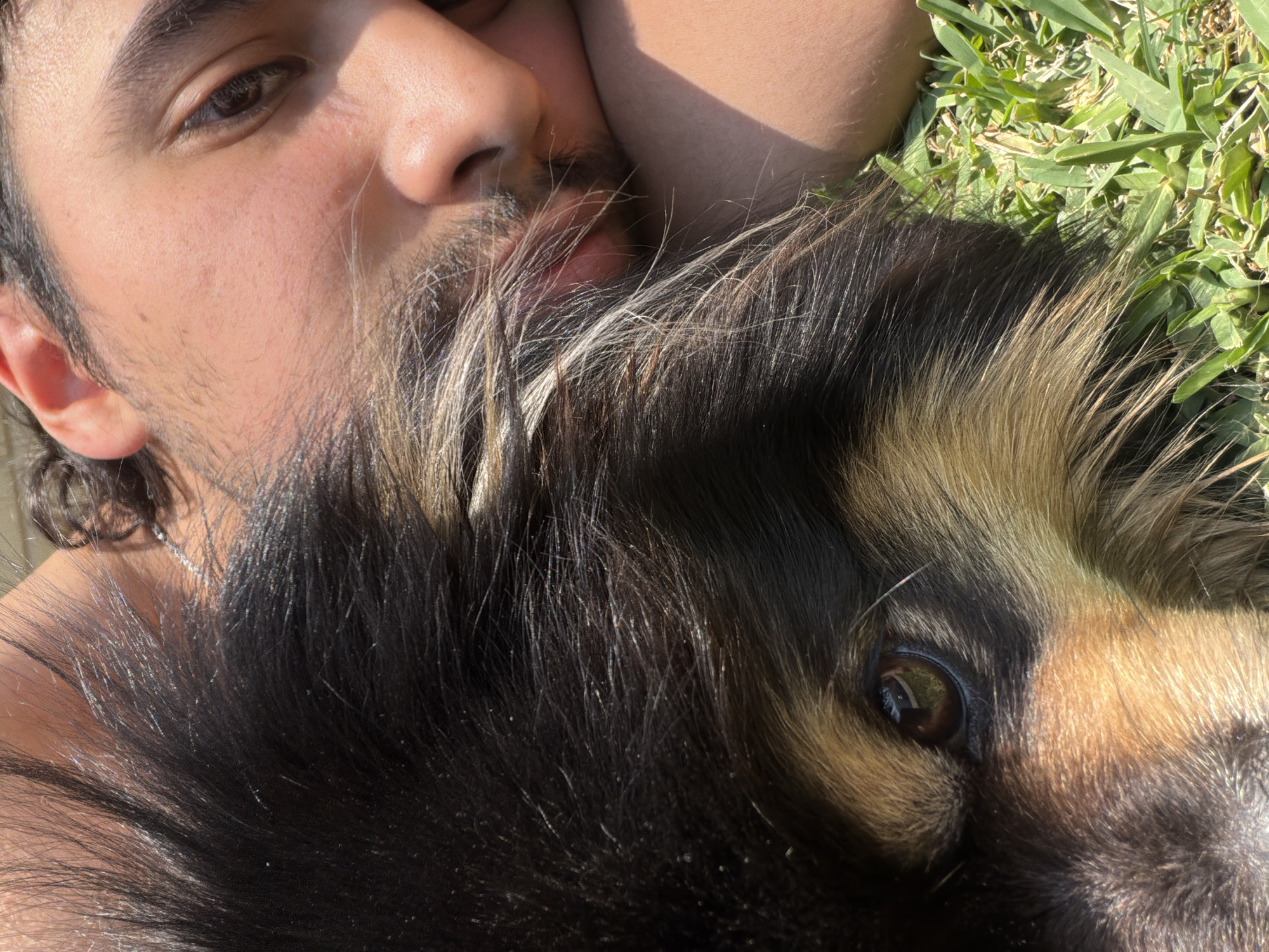 A close-up, high-angle shot shows a man lying down with a dog, possibly outside on a lawn. The man's face is partially visible, with his eye open and looking directly at the camera. He has dark hair, a stubbly beard, and fair skin. Part of his arm rests above his head, which is close to the dog's fur. The dog, primarily black with tan markings around the face, has one eye visible, also looking at the camera. The background consists of green grass. There is no information to confirm this image was taken in Cuautla, Mexico.