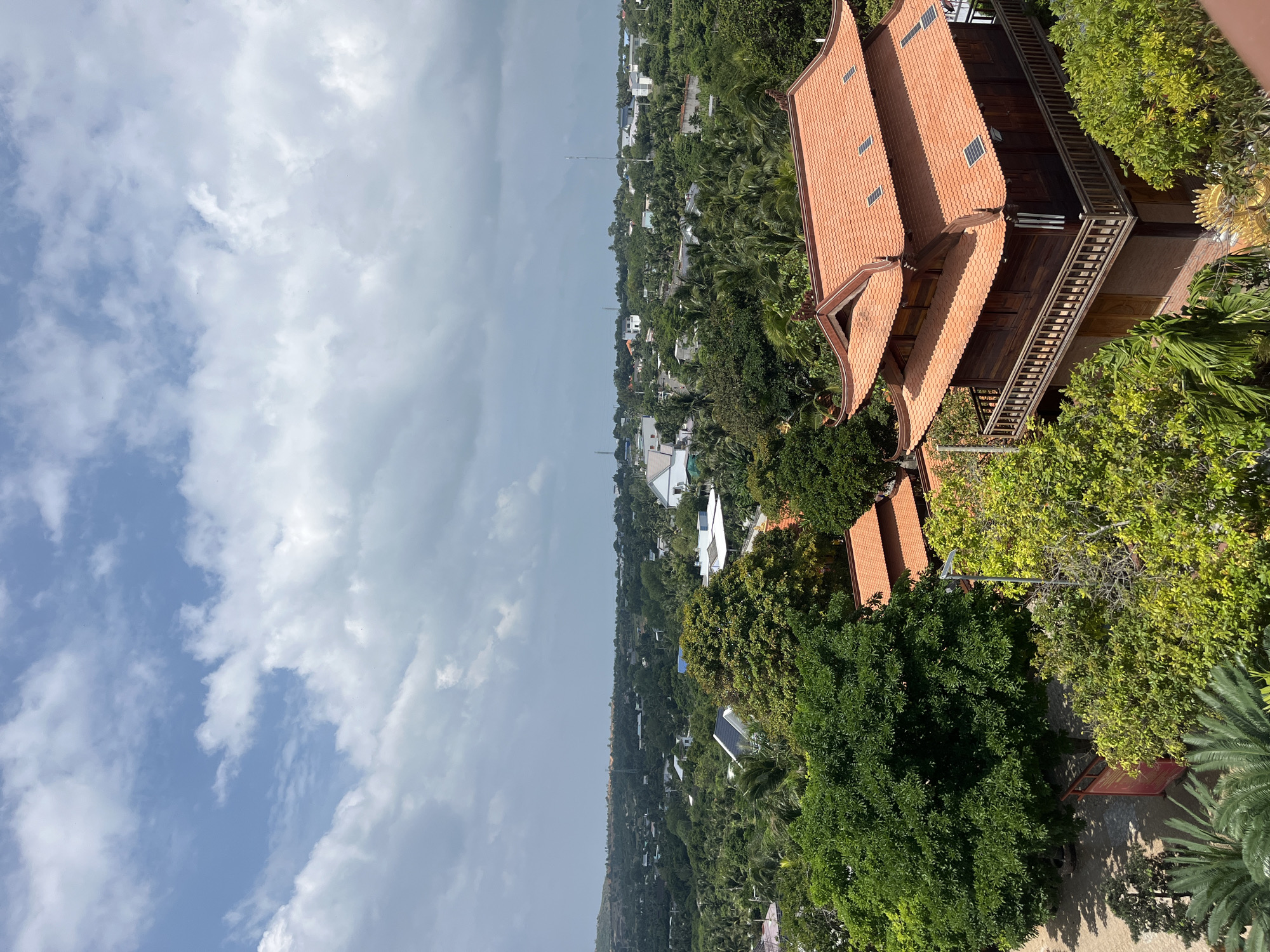 The image captures an elevated view of a village or town nestled amidst lush greenery, likely in Mũi Né, Vietnam. In the foreground, a traditional Vietnamese-style building with an orange tiled roof and wooden exterior stands prominently. The building features multiple tiers, a wrap-around balcony with wooden railings, and numerous windows. Surrounding the structure are various trees and palm fronds, suggesting a tropical or subtropical environment.
Beyond the immediate foreground, the landscape unfolds into a dense expanse of trees, interspersed with houses and structures. The architecture of these buildings appears to be a mix of modern and traditional styles, with white walls and tiled roofs being common. A hint of the coastline or a distant body of water can be inferred from the hazy horizon.
The sky above is partly cloudy, with a combination of bright blue patches and large, fluffy white clouds, indicating fair weather with potential for scattered cloud cover. The overall impression is one of a tranquil, verdant community, likely a resort town or a residential area with significant natural foliage. No people are visible in the image, and there are no discernible actions or interactions taking place. The scene is static and peaceful, focusing on the architecture and the natural environment.