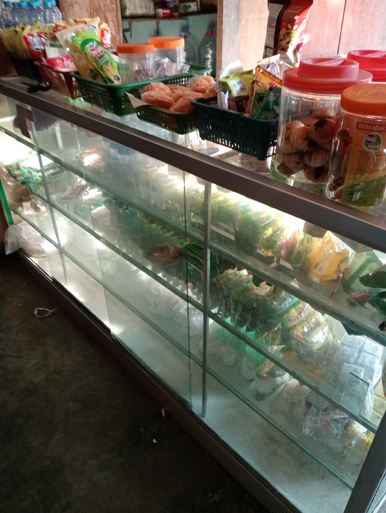 The image shows a glass display case filled with various food items and snacks, likely in a small shop or market in Grabag, Indonesia. The display case is made of glass and metal framing, with multiple shelves. On the top shelf, several plastic containers with orange lids are visible, one filled with round, brown baked goods. Next to these are green baskets containing packaged snacks. The shelves below are filled with numerous individually packaged food items, mostly in colorful plastic wrappers, including what appear to be instant noodles, crackers, and other processed snacks. Some items are also visible in clear plastic bags. The overall impression is of a well-stocked retail environment. The background suggests a simple interior with wooden elements and other shelves stocked with goods, though they are out of focus. The lighting appears to be artificial, casting a yellowish hue on some of the items.