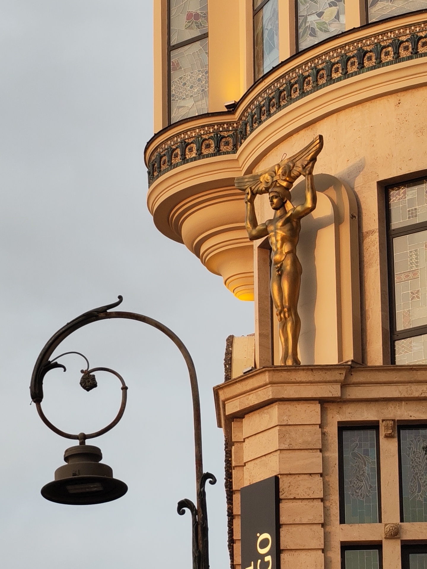 The image captures a segment of an ornate building in Batumi, Georgia, under a pale sky. The building's facade is constructed from light-colored stone blocks, with a section of a rounded balcony featuring decorative metalwork in shades of green and gold. Above the balcony, a statue of a golden-skinned, muscular male figure stands in a niche, holding a winged object above his head. The statue is illuminated by warm, angled light, likely from the setting sun. Stained-glass windows with intricate patterns are visible on the upper levels of the building, and a modern black sign with gold lettering is partially visible at the lower right. In the foreground to the left, a dark, wrought-iron lamppost with a scrolled design curves over the scene, its base and light fixture dark and detailed. The overall impression is one of architectural grandeur with artistic embellishments.