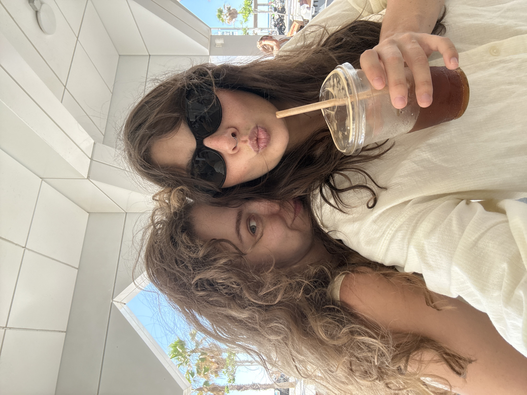 Two women are taking a selfie outdoors, possibly at a cafe in Tel-Aviv, Israel, with palm trees and a clear blue sky visible in the background. The woman on the right is wearing sunglasses and has her lips pursed, holding a cold drink with a straw. The woman on the left is leaning into the frame, smiling. They appear to be casually dressed. The scene suggests a relaxed atmosphere, likely during daylight hours.