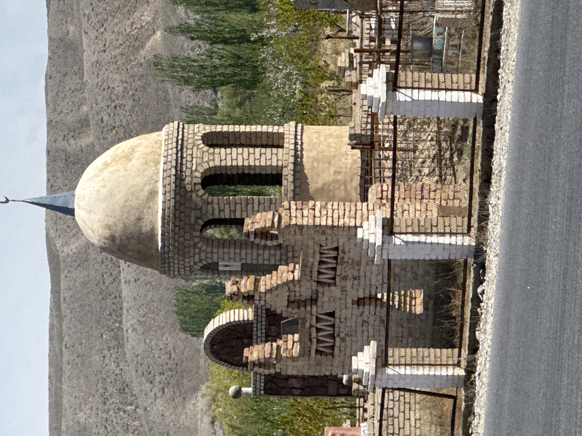 The image, when viewed as if correctly oriented, presents a light-colored brick structure, likely a mausoleum or memorial, situated in a mountainous, arid landscape. The building features a prominent, smooth, light-beige dome at its apex. Extending from the dome's side is a slender, pointed spire adorned with a crescent moon finial, hinting at Islamic architectural influence.
The structure itself is built from light-toned bricks, arranged in decorative patterns. It includes several arched openings or windows in its upper section and more intricate brickwork in its lower parts, featuring stepped elements and smaller arches. Some sections, possibly pillars or railings, are painted white, providing a contrast to the brick. Metal fencing surrounds the structure in places, suggesting an enclosed boundary.
The setting is characterized by sparse green trees and shrubs, some with lighter foliage, contrasting with the barren, rocky mountains that dominate the background. A paved or gravel road runs along the immediate foreground of the scene. The bright, clear sky and short shadows indicate strong daylight, likely midday. No people or activities are visible in the image. There is no visible text. The overall scene is consistent with the landscape and architectural styles found in Central Asia, such as Kara-Talaa, Kyrgyzstan.