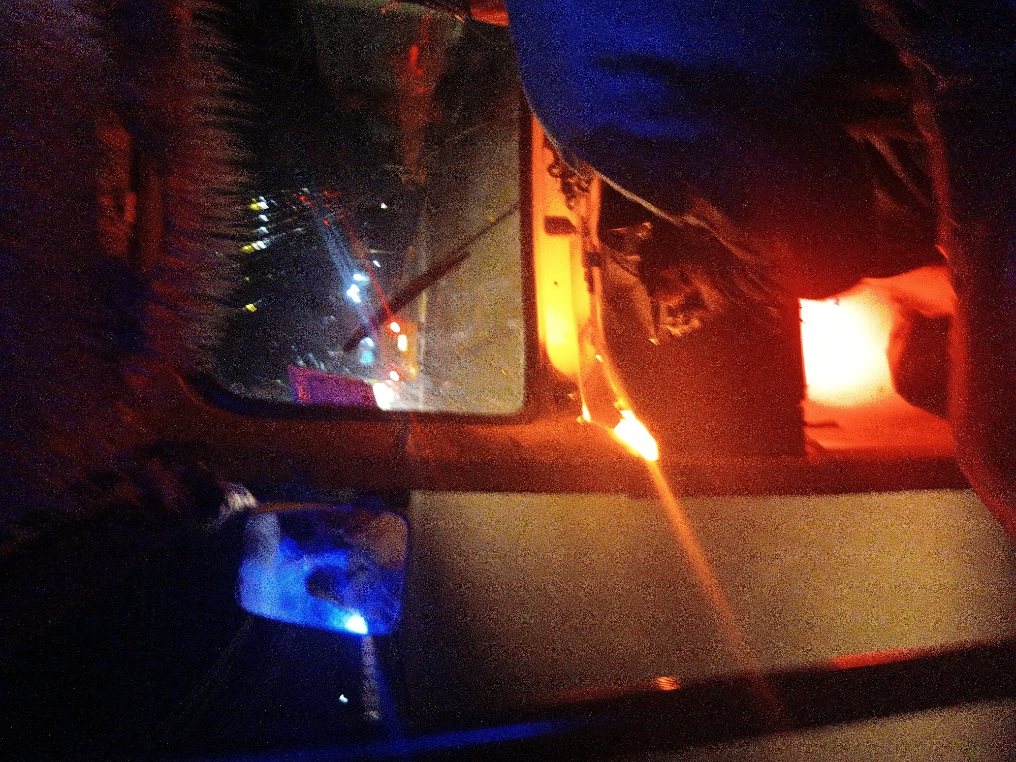 The image depicts the interior of a vehicle at night, likely a tuk-tuk, with a driver visible in the foreground. The driver, wearing a blue garment, is partially obscuring the steering wheel and dashboard area. The exterior environment is characterized by darkness punctuated by various light sources, including headlights from other vehicles on a road. The foreground of the interior is illuminated by an orange glow, suggesting interior lighting or reflections from external sources. The roof of the vehicle appears to be adorned with thatch or a similar material. The scene implies movement through an urban setting at night, consistent with a street in Jalingo, Nigeria.