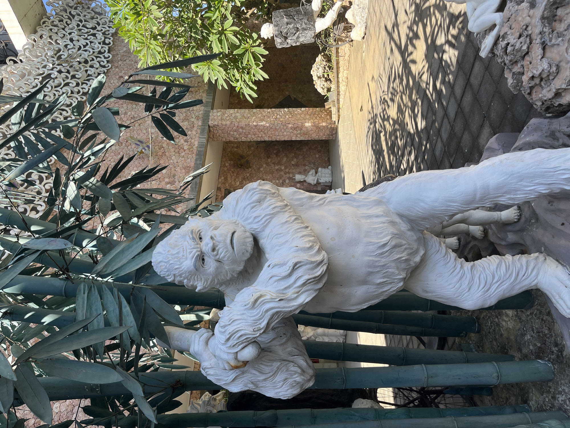 The image depicts a white statue of a gorilla standing next to artificial bamboo stalks in Sukawati, Indonesia. The gorilla statue is detailed with fur and has a stern expression. Behind the gorilla, a building with a textured stone facade and white decorative elements is visible. Lush green foliage frames the scene, with sunlight casting dappled shadows on the paved ground. Other statues, including what appear to be human figures, are scattered around the area. The overall setting suggests an outdoor decorative space or garden.