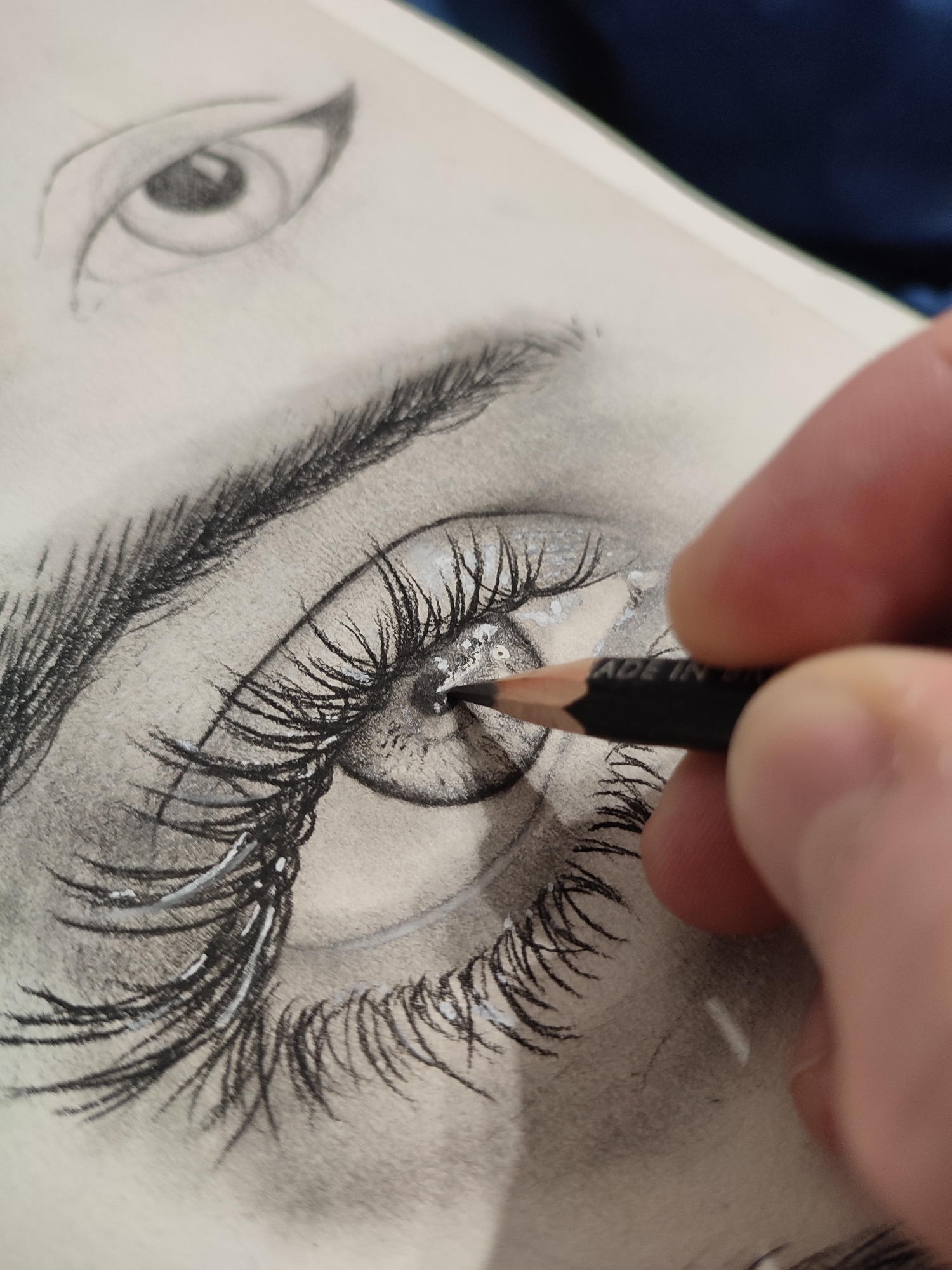 A close-up, overhead, extreme-angle shot shows a person's hand holding a dark pencil with the writing "MADE IN UNKNO..." visible on its side, actively drawing on a piece of paper. The drawing is a realistic sketch of a human eye with detailed eyelashes. The background is blurred and appears to be a dark blue fabric. The paper has a textured surface and is slightly off-white. The lighting is diffused, highlighting the texture of the paper and the pencil strokes.