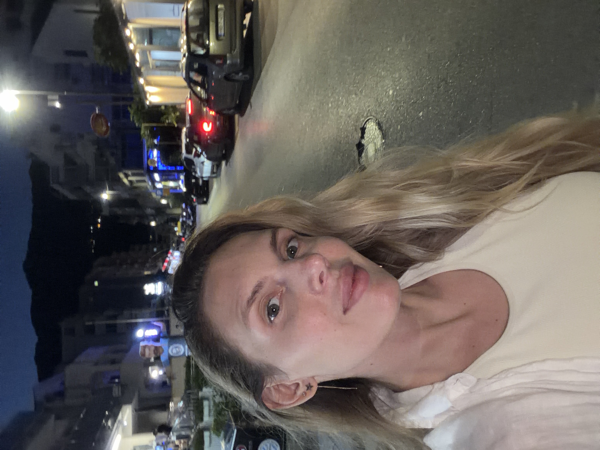 A woman with long blonde hair and light skin is taking a selfie at night on a street in Budva, Montenegro. She is wearing a white shirt and a cream-colored top. Her left ear is visible, adorned with a star-shaped earring connected to a dangling chain. In the background, a street is lined with buildings and cars. The street is illuminated by streetlights and the headlights of vehicles. The overall scene depicts a nighttime urban environment.