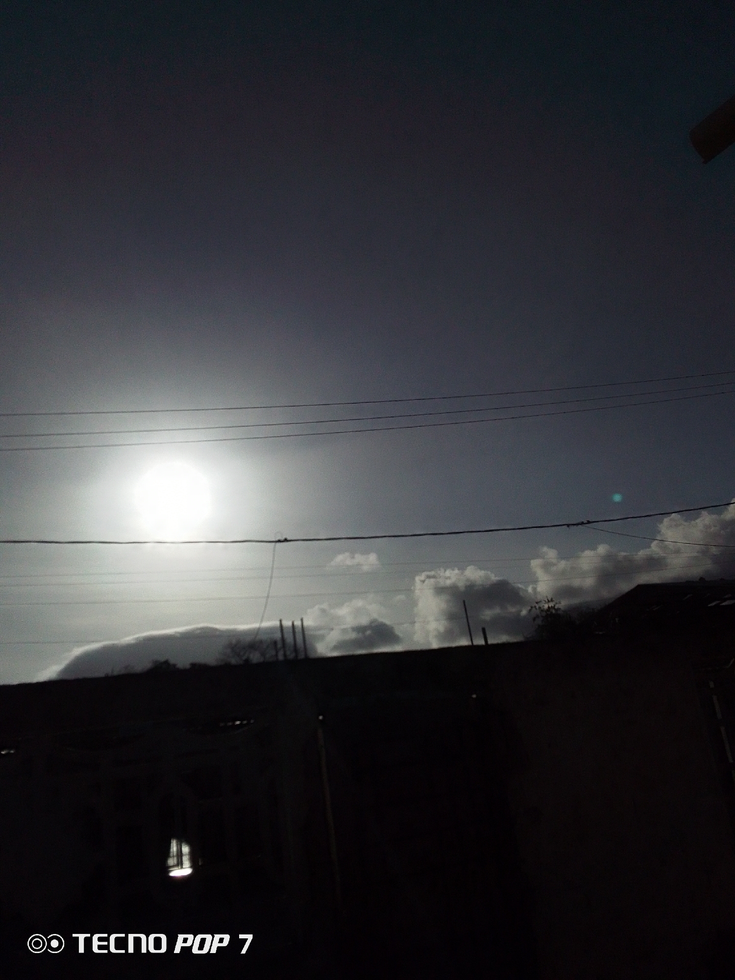 The image shows a view of the sky with the sun obscured by clouds, taken in Altagracia, Venezuela. Power lines are visible across the sky. Below the horizon, there is a dark silhouette of buildings. The image was taken with a Tecno Pop 7 phone.