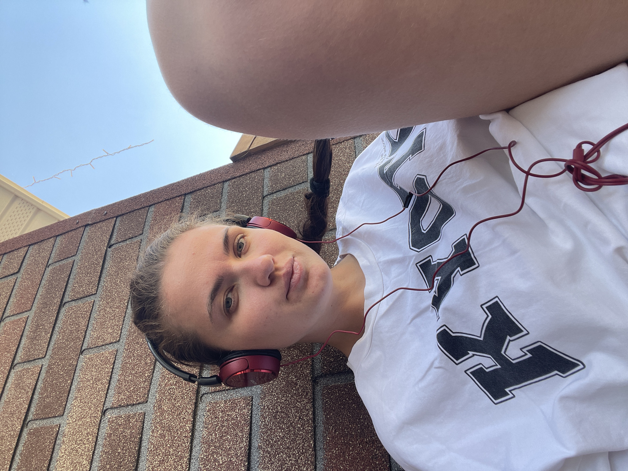A young woman with light skin and brown hair in a braid is lying on a roof covered in brown asphalt shingles. She is wearing red headphones and a white t-shirt with black lettering. A section of the sky is visible in the background. The location appears to be in a residential area, possibly the city of Мосметаллоконструкция Онт, Russia, as indicated.