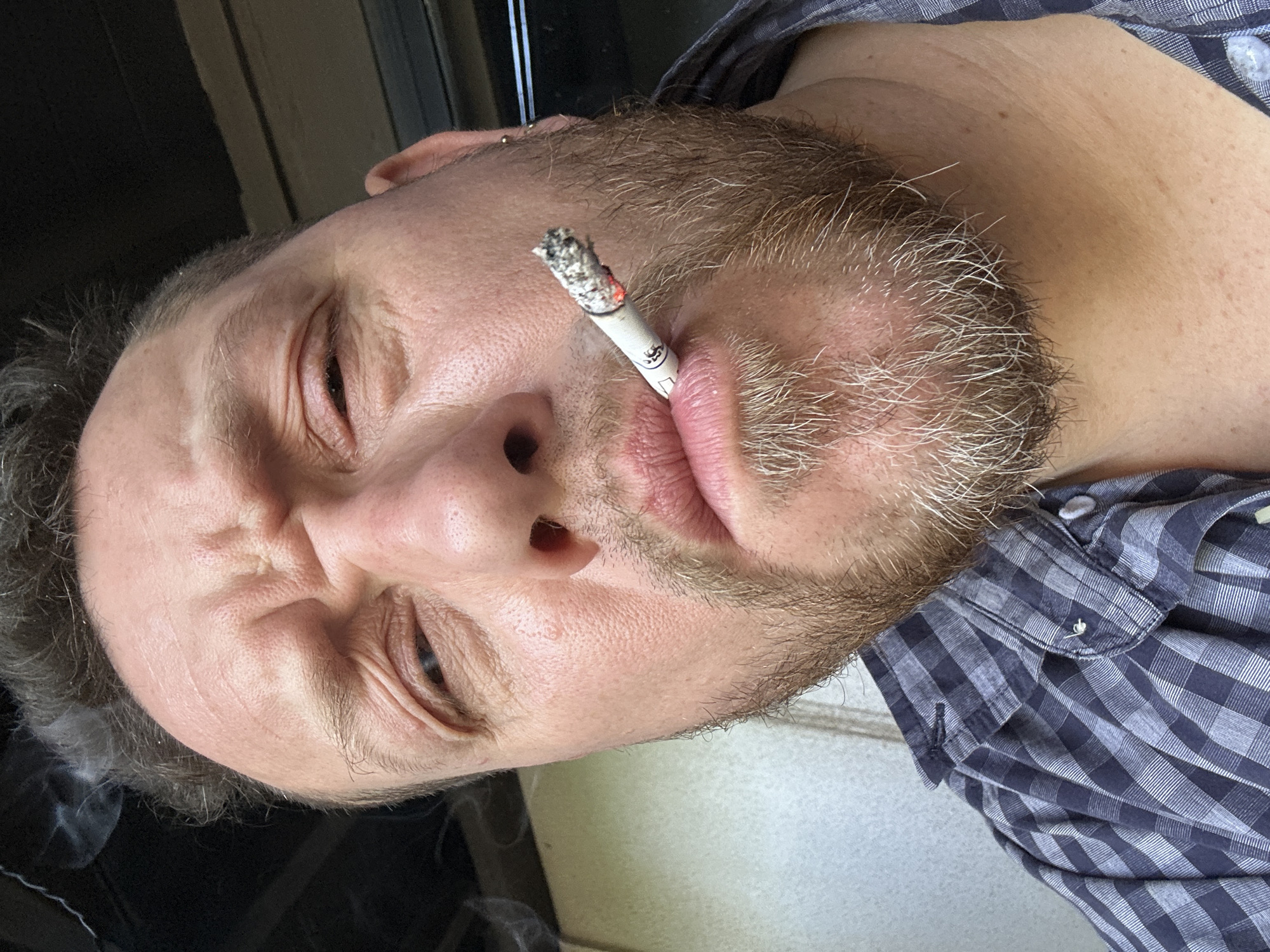 A man with a light complexion and a beard with graying highlights is depicted in the image. He is holding a lit cigarette in his mouth. He is wearing a blue and white checkered shirt. The photograph seems to have been taken in Новоглаголево, Russia.