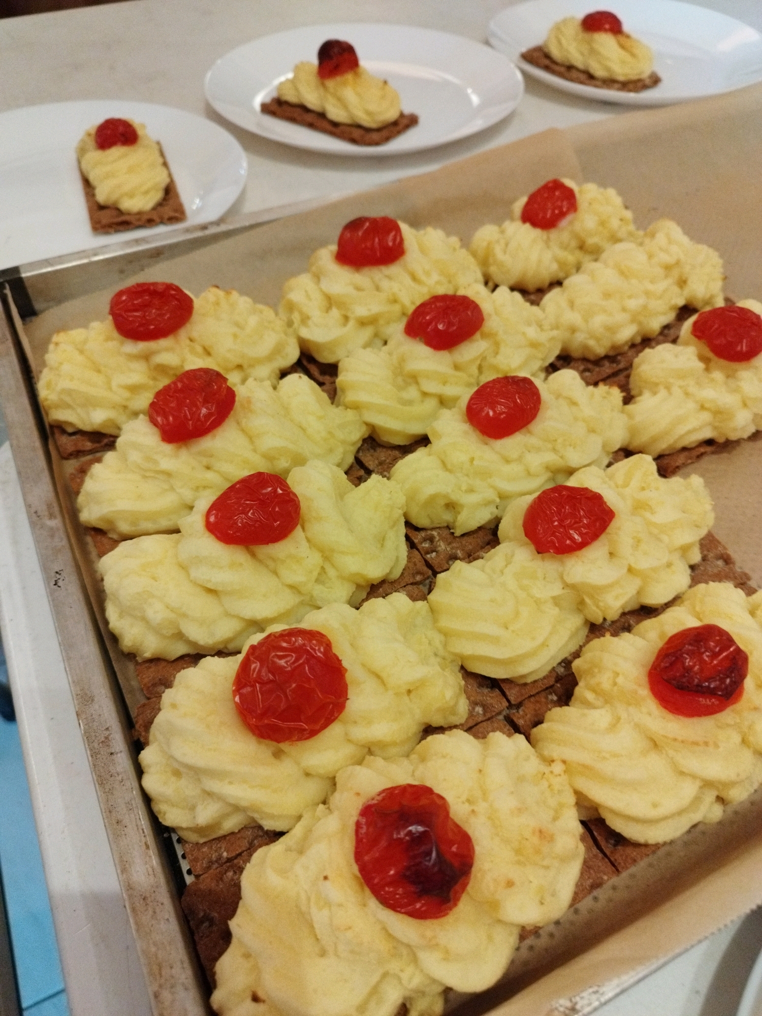 A tray of individually prepared potato and tomato appetizers sits on a counter. Two similar appetizers on white plates are in the background. The appetizers are composed of a rectangular base of dark bread topped with piped mashed potatoes and a single, halved cherry tomato. The tray is on a light-colored surface, possibly a kitchen counter or table. The lighting suggests an indoor setting. The scene is forensic, matter-of-fact, and devoid of any specific location information within Bucharest, Romania, or any indications of people or actions.