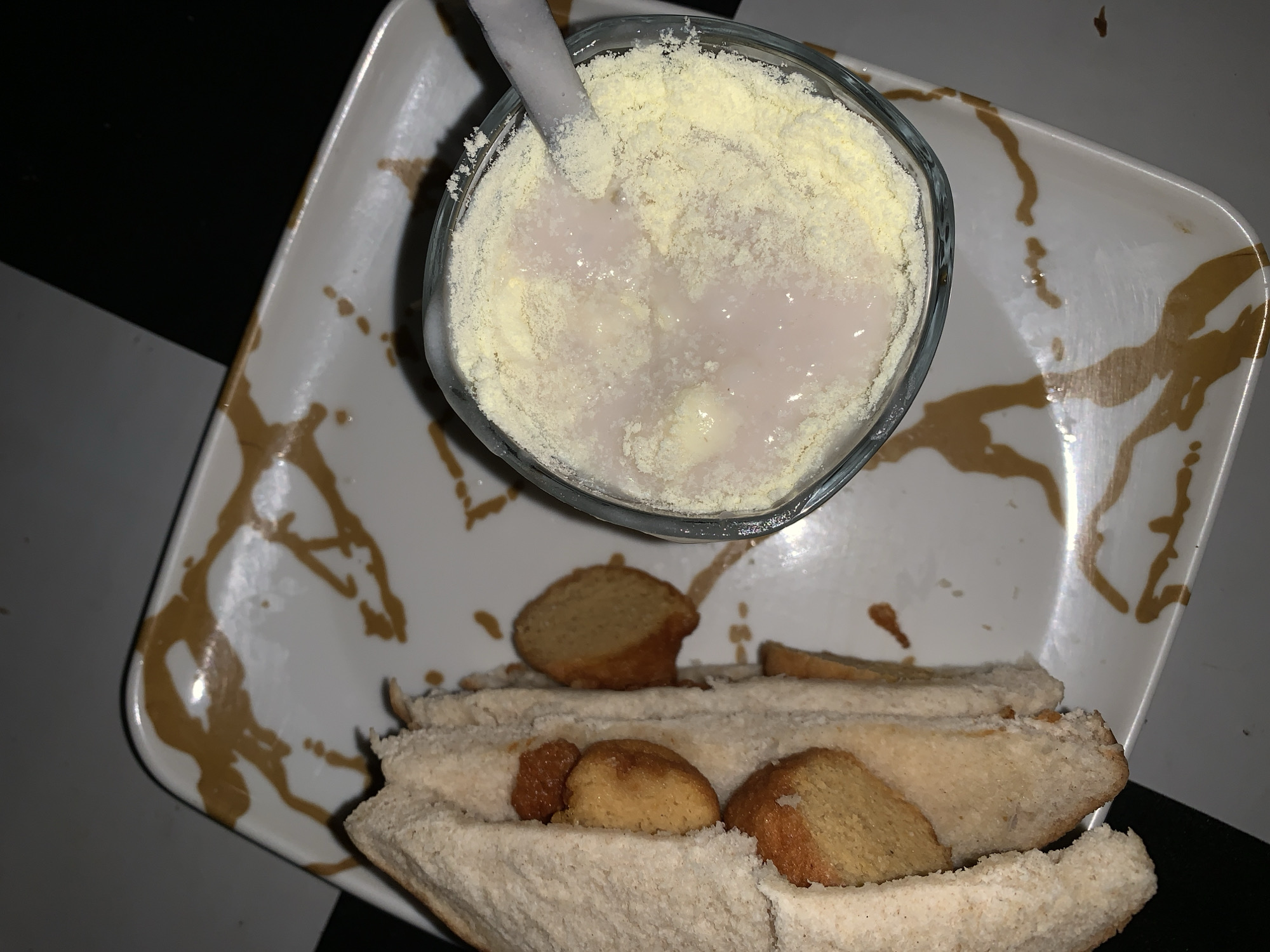 A top-down view shows a white rectangular plate with gold splatter marks. On the plate are the following items:
1. A clear glass containing a white liquid and a thick layer of yellow powder. A white spoon is inserted into the mixture.
2. A white bread sandwich, cut into multiple pieces, with what appears to be small, round, golden-brown cookies or pastries nestled within the bread.
The background is a black and white checkered surface. It is not possible to identify the city or location with just the image.