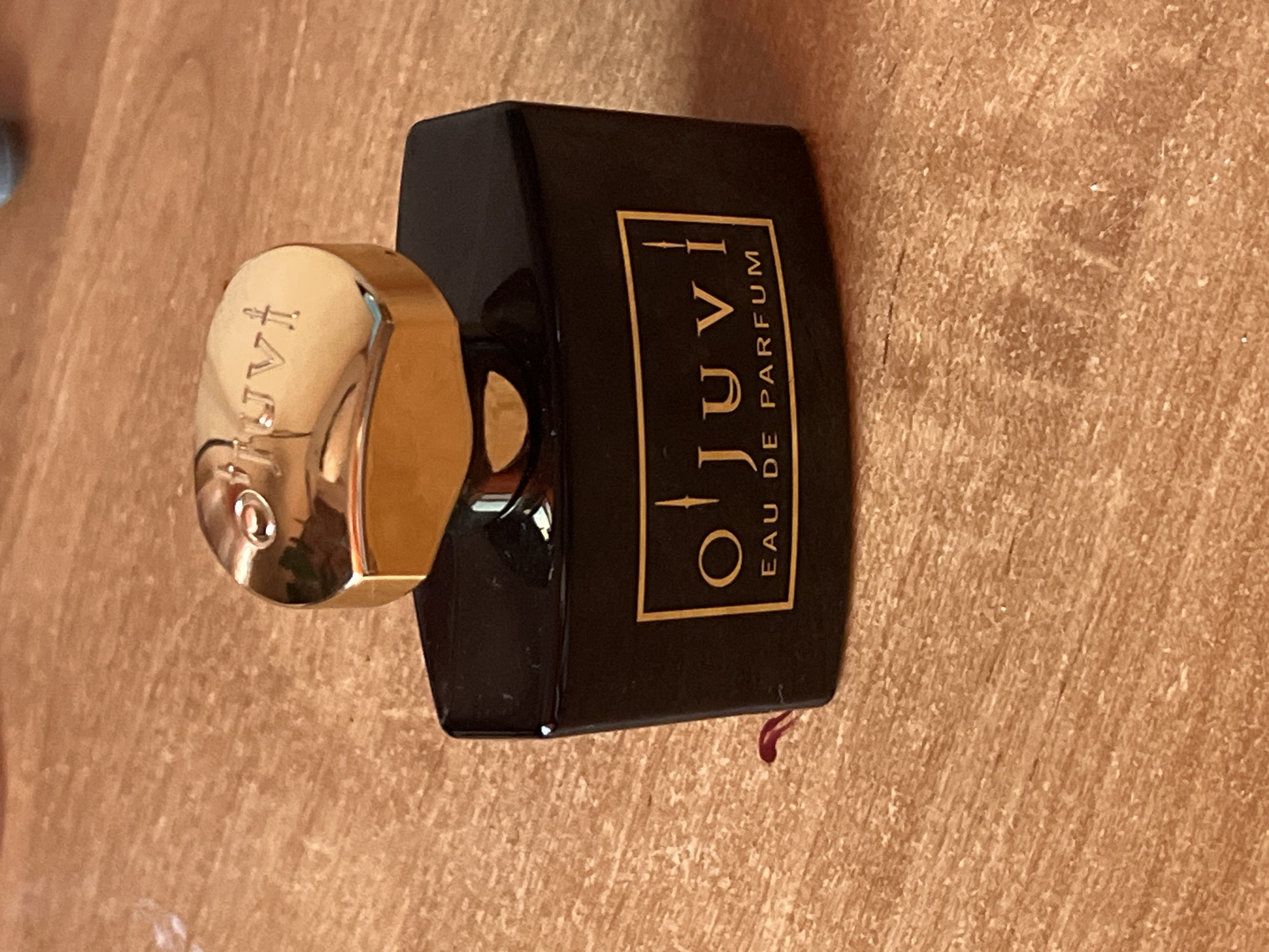 A black, hexagonal perfume bottle with a gold cap sits on a wooden surface. The bottle has a gold label with the text "O'juvi EAU DE PARFUM" in a stylized font. The cap has the word "O'juvi" embossed on it. There is a small red stain on the wooden surface to the left of the bottle.