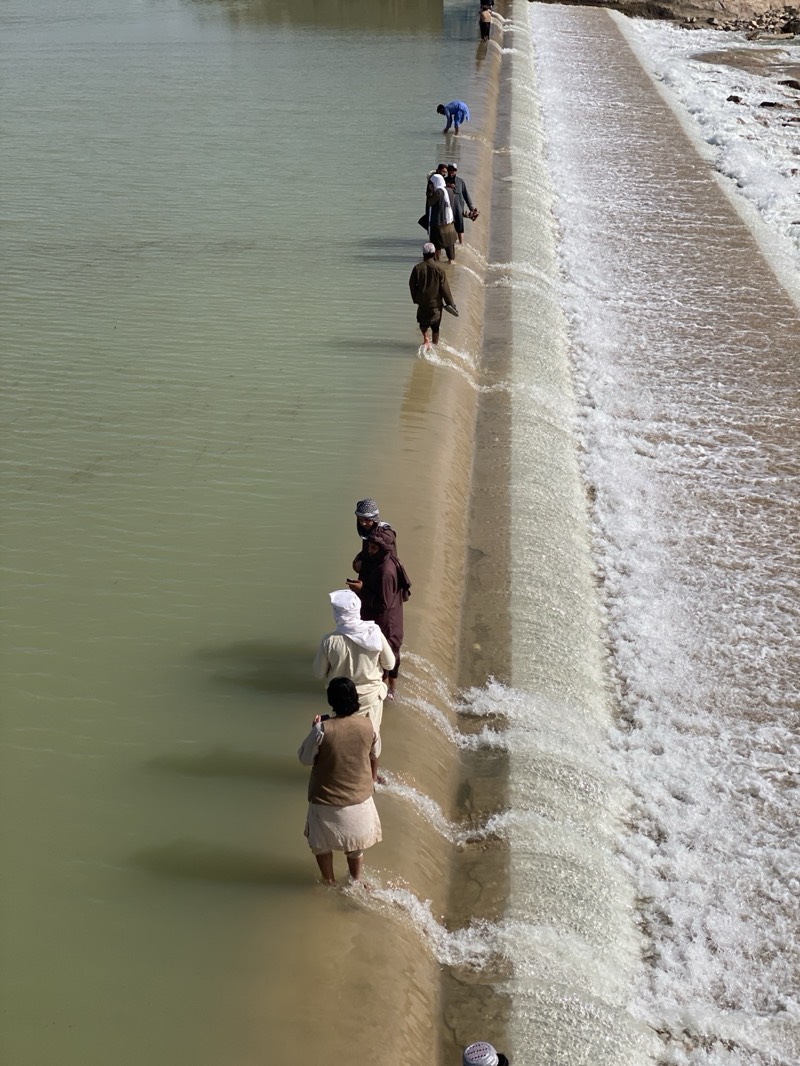 A high-angle, long-shot photograph shows a line of several men crossing a dam in what appears to be a river. The location is in the city of Kim, Afghanistan. The water is a muddy green color and flows from left to right. The men are wading through the water as they walk along the dam's edge, some are dressed in traditional Afghan attire, including long robes and head coverings. The sky is not visible, but the light suggests a bright, sunny day. The dam is made of concrete and has water flowing over its edge.