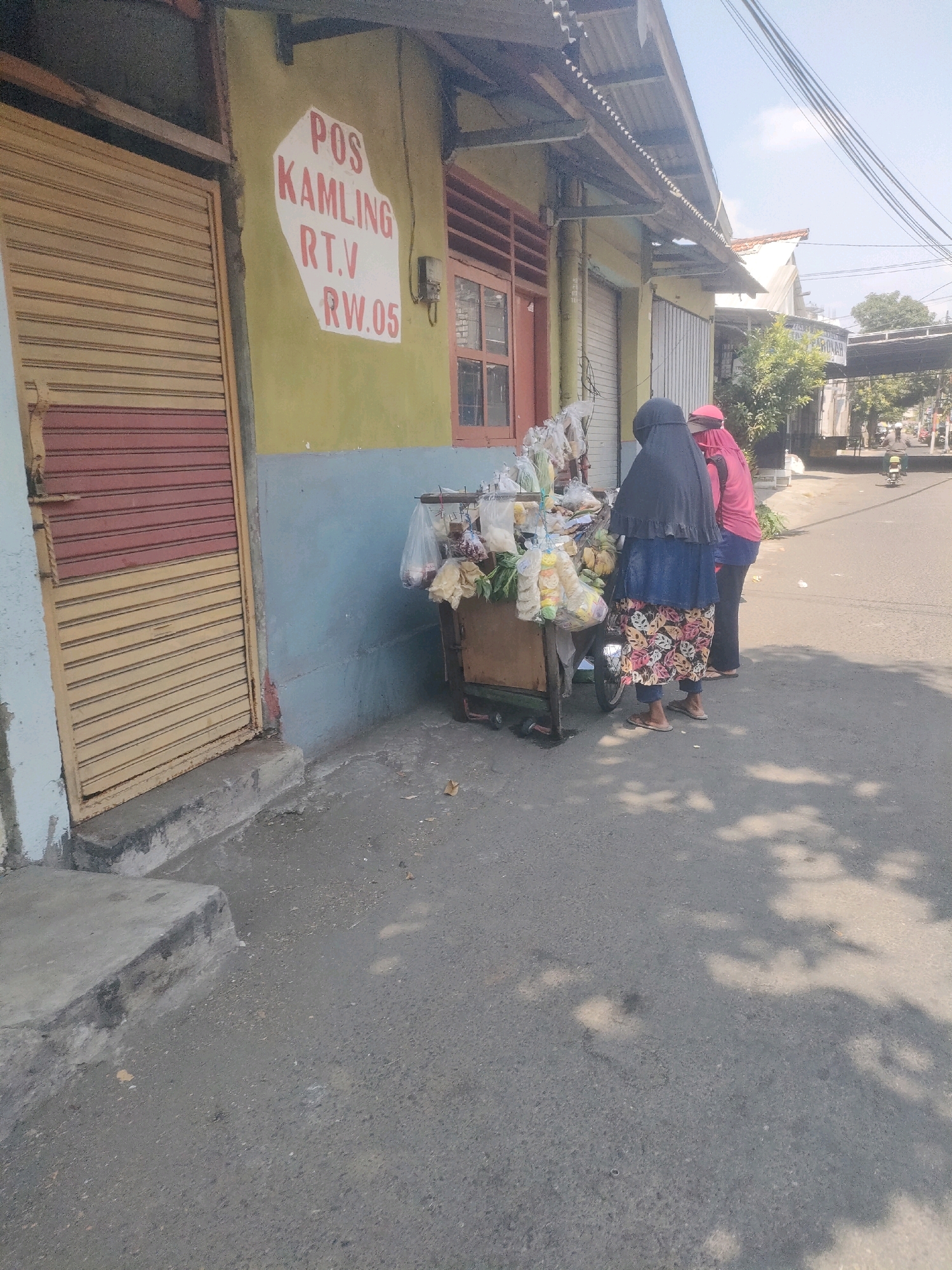 The image depicts a street scene in Surabaya, Indonesia, with a particular focus on a roadside stall and its patrons. A yellow and green building with a corrugated metal roof stands to the left, featuring a rolled-down metal shutter door. A sign on the building reads "POS KAMPLING R.T.V RW.05" in red lettering on a white background. Adjacent to the building is a mobile cart laden with various items, including packaged snacks and what appear to be fruits or vegetables. Two individuals are standing by the cart, seemingly engaged in a transaction or browsing the goods. One person wears a black hijab and a blue skirt with a floral pattern, while the other is in a pink headscarf and pink top. The ground is paved with asphalt, and sunlight casts shadows from unseen trees onto the surface. In the background, a road stretches into the distance, with more buildings and trees visible. A motorcycle and a few other vehicles are present on the road, suggesting a moderate level of activity. The overall impression is of a typical urban street scene in a residential or commercial area of Surabaya.