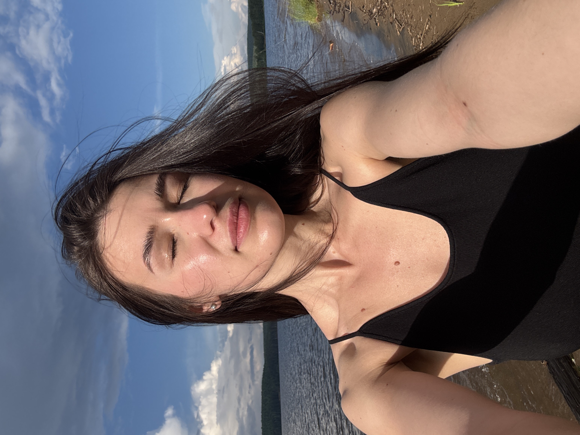 A young woman with long, dark hair is taking a selfie outdoors. She is wearing a black tank top. Her eyes are closed, and her face is tilted slightly upwards, catching the sunlight. Her skin appears to be glowing with a light sheen. She is positioned in front of a body of water, likely a lake, with a shoreline visible to her right. Beyond the water, a treeline forms the horizon. The sky above is blue with scattered clouds. This scene is consistent with a location on the Kama River near Chaikovsky, Russia.